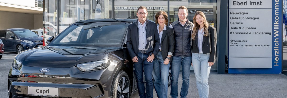 Eine Gruppe von vier Personen steht neben einem schwarzen Ford Capri in einem Autohaus. Hintergrund zeigt das Geschäftsschild.