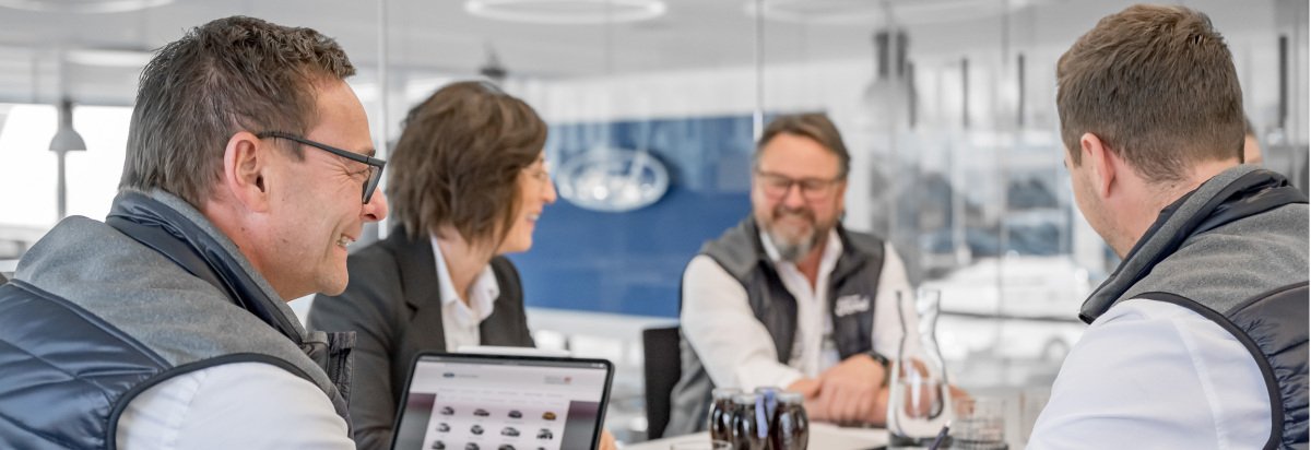 Geschäftsbesprechung mit vier Personen in einem modernen Büro, Ford-Logo im Hintergrund, Laptops und Getränke auf dem Tisch.