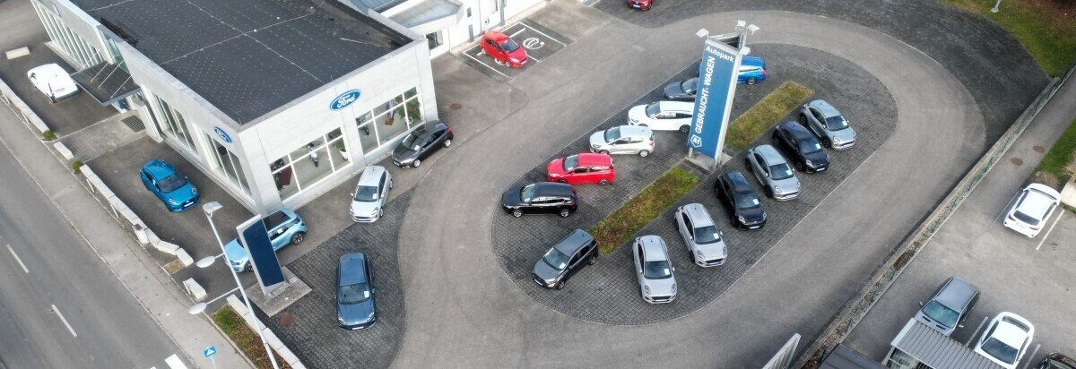 Luftaufnahme eines Autohauses mit mehreren Autos auf dem Parkplatz und dem Verkaufsgebäude im Hintergrund.