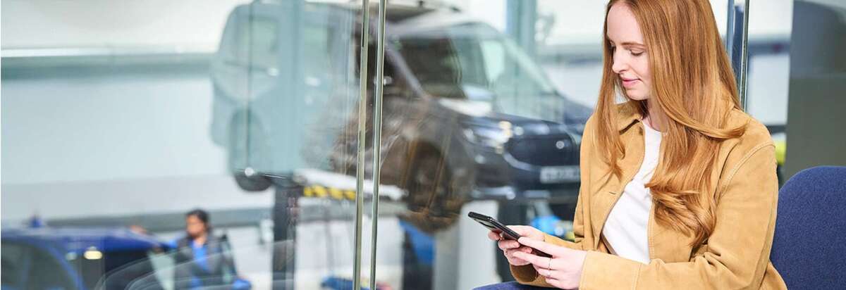 Eine Frau mit langen roten Haaren sitzt in einer Werkstatt und schaut auf ihr Smartphone, während ein Auto im Hintergrund repariert wird.