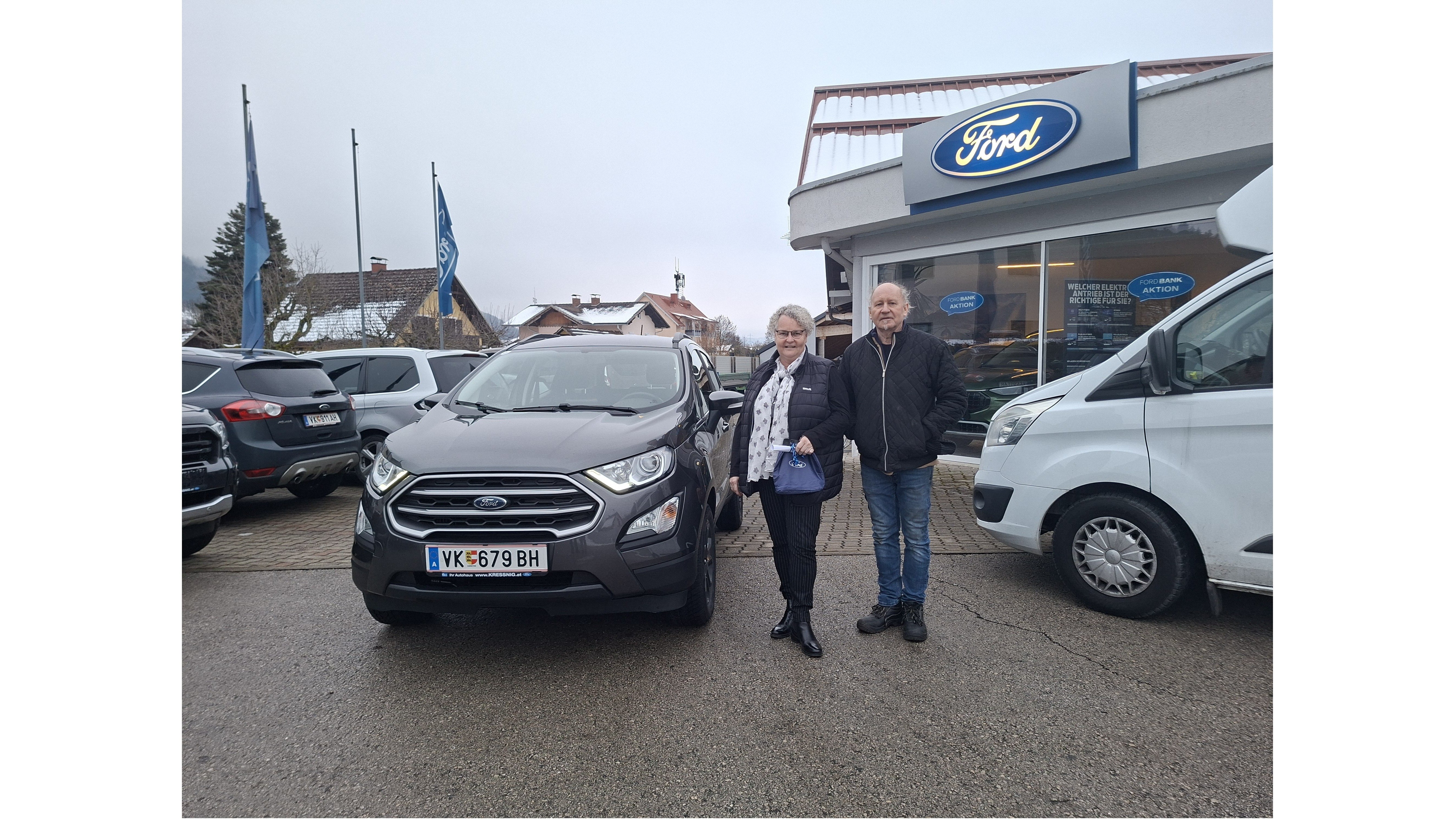 Ein Paar steht neben einem grauen Ford EcoSport vor einem Ford-Händler. Wolkiger Himmel im Hintergrund.