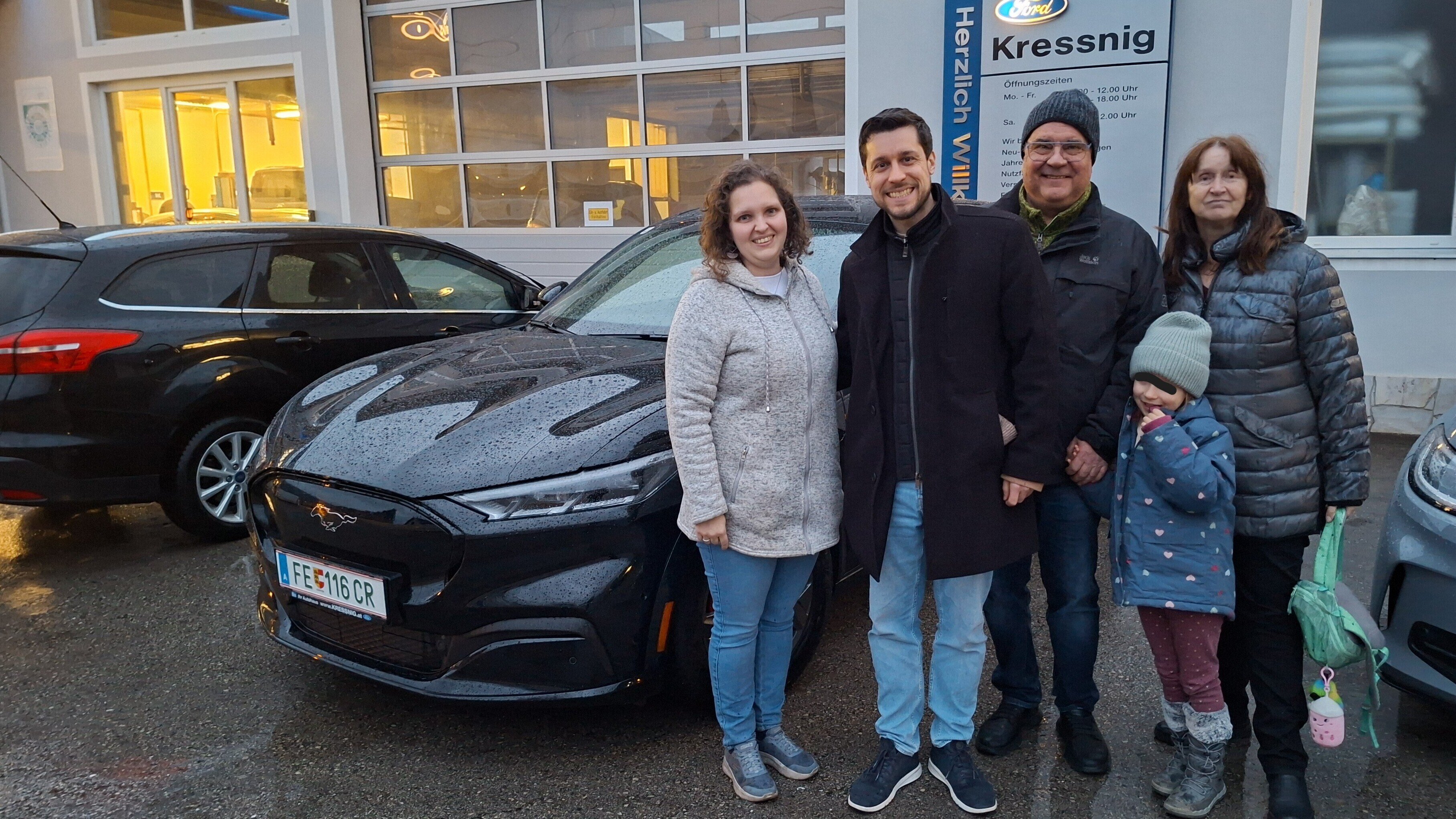 Eine Familie steht lächelnd neben einem schwarzen Ford Mustang, neben Autos in einer Garage bei Kressnig.
