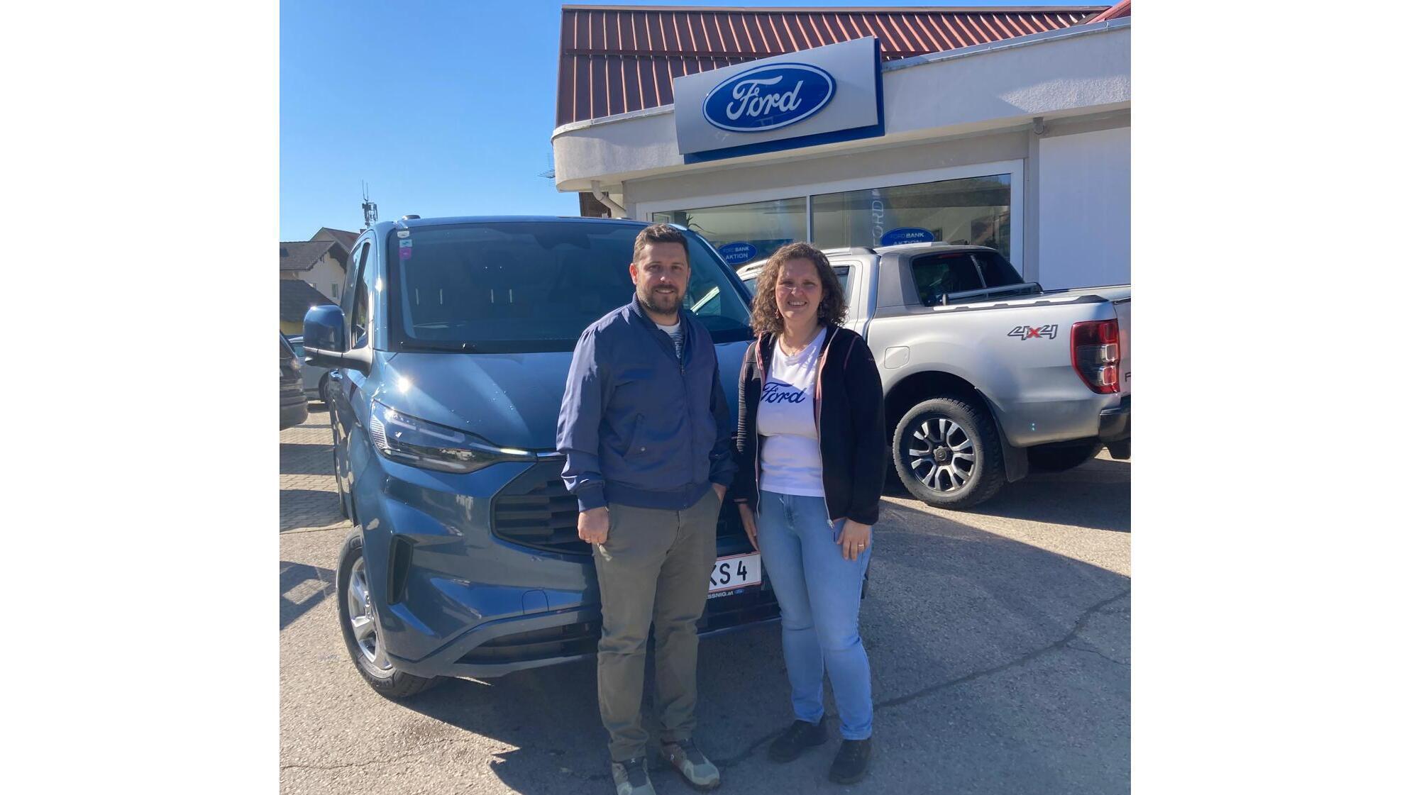 Ein Mann und eine Frau stehen vor einem neuen Ford-Fahrzeug bei einem Autohaus mit Ford-Schild.