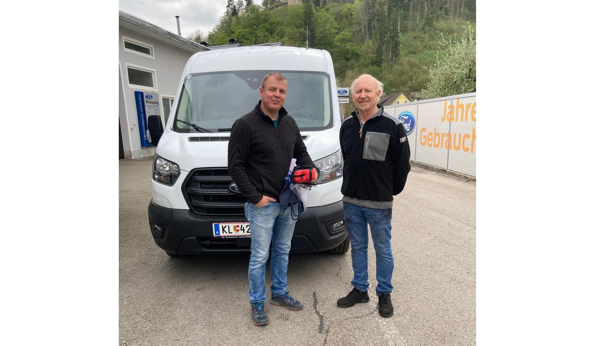 Zwei Männer stehen vor einem weißen Lieferwagen, umgeben von grüner Natur und einer Werkstatt.