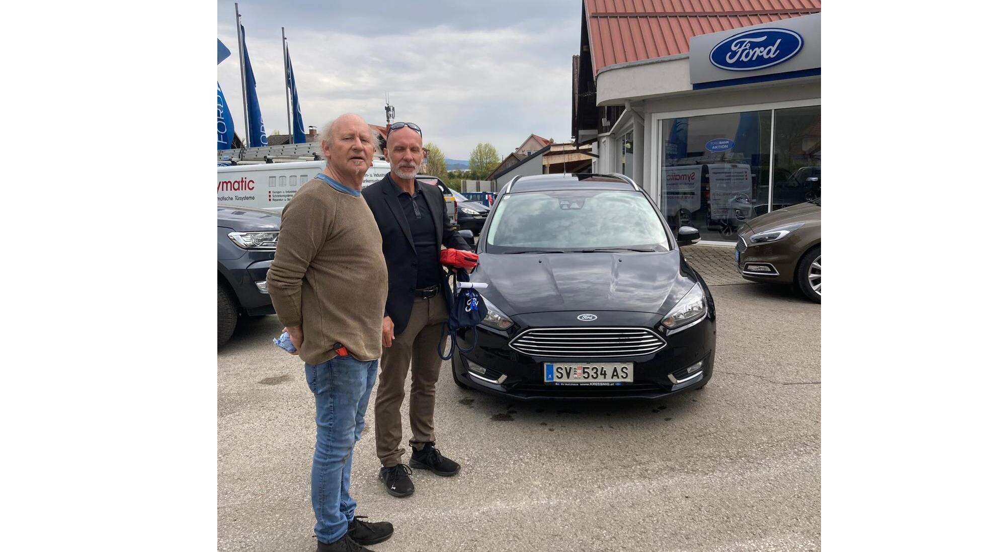 Zwei Männer stehen vor einem schwarzen Ford am Autohaus, hinter ihnen das Ford-Schild und andere Fahrzeuge.