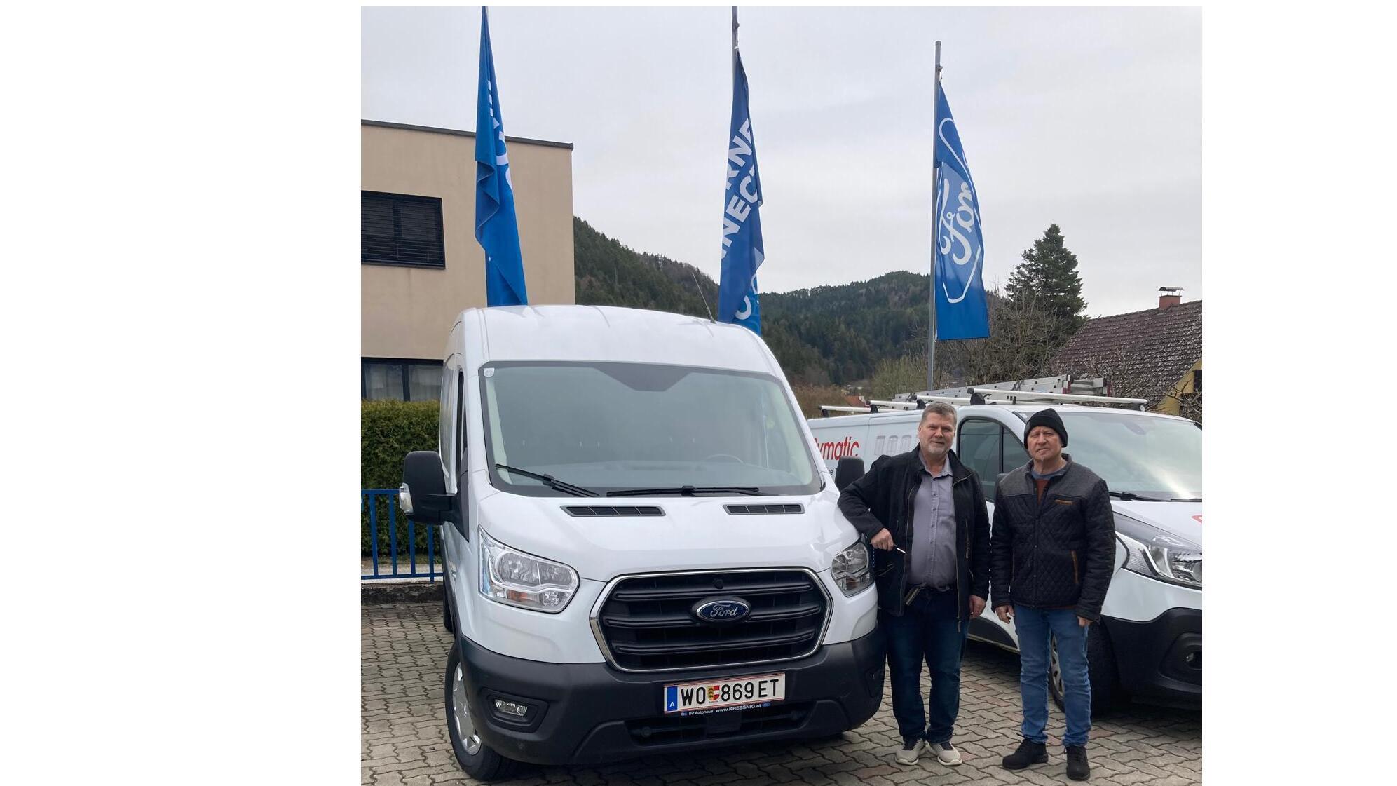Zwei Männer stehen vor einem weißen Ford Transporter mit Fahnen im Hintergrund, in einer ländlichen Umgebung.