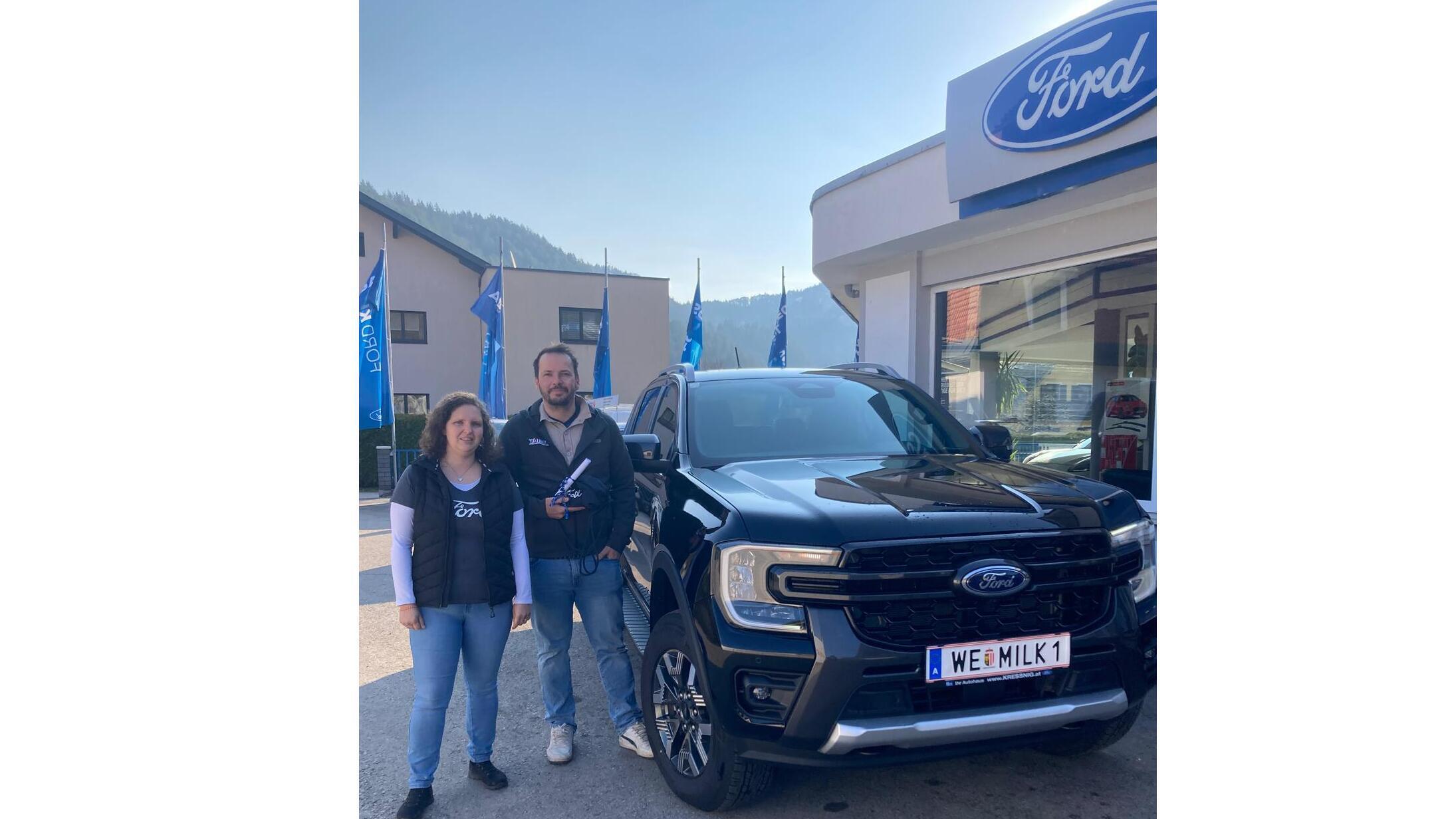 Zwei Personen stehen vor einem neuen Ford Fahrzeug in einem Autohaus, umgeben von Flaggen und Bergen.