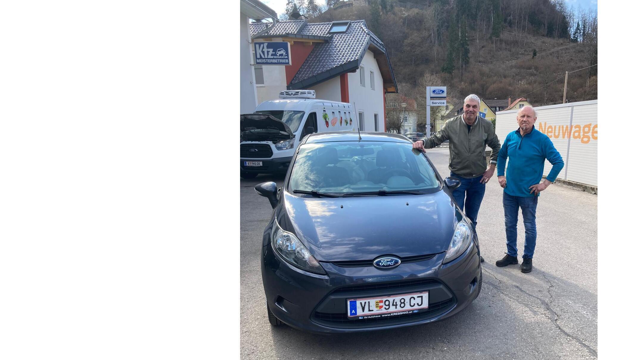 Zwei Männer neben einem grauen Ford Fiesta vor einem Autohaus in ländlicher Umgebung.