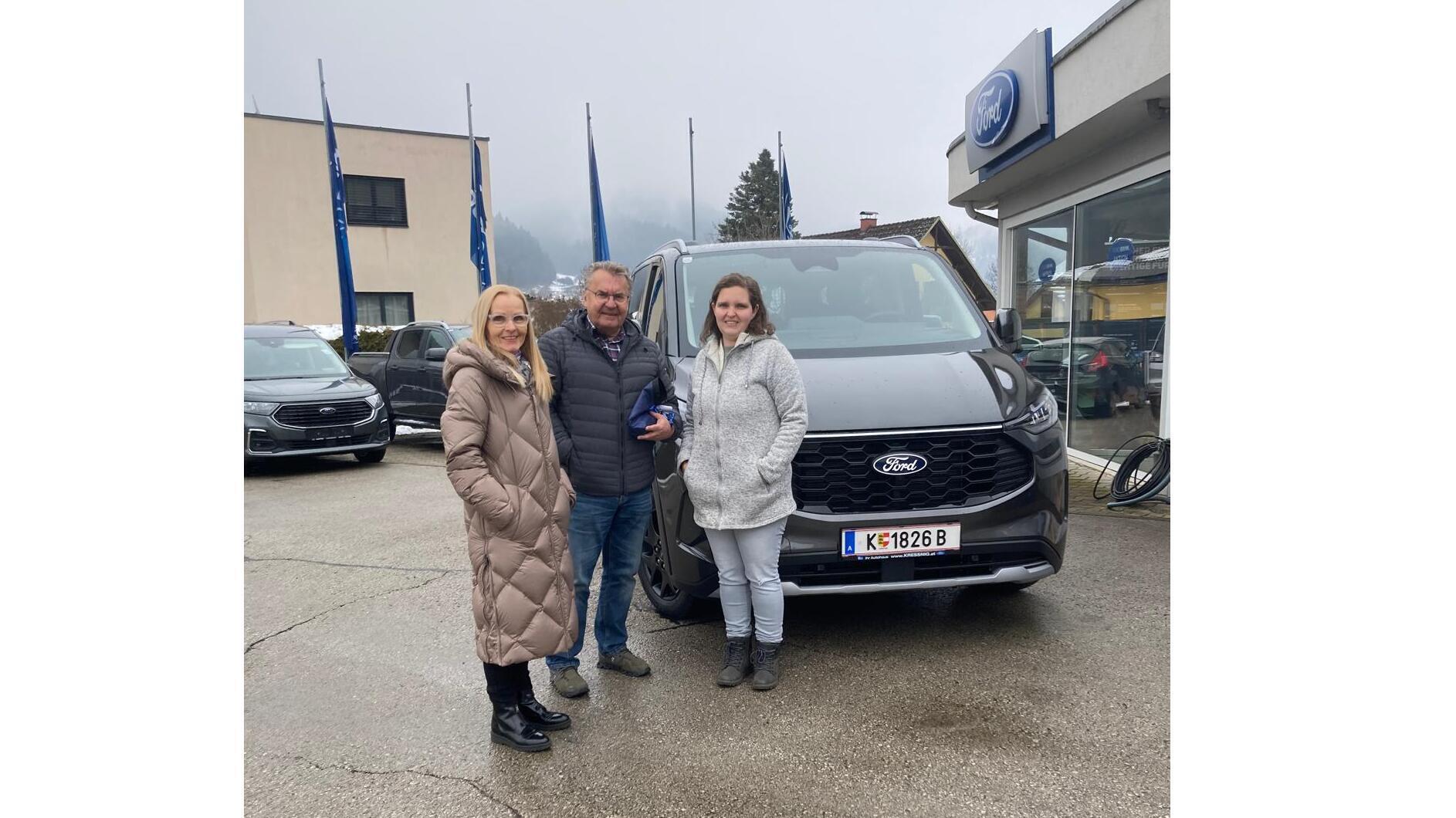 Drei Personen stehen vor einem neuen Ford-Auto in einem Autohaus, umgeben von Flaggen und Nebel.