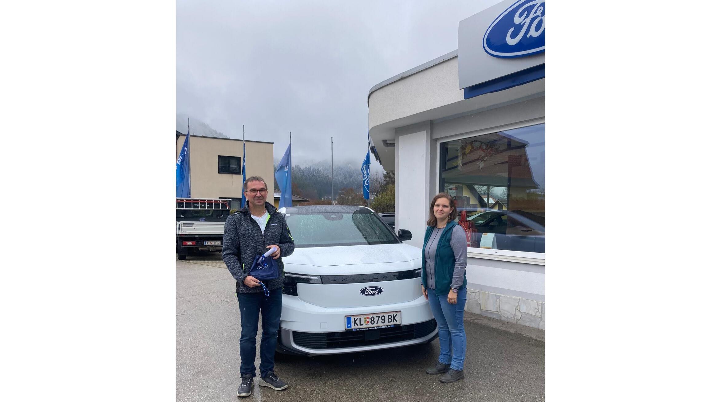 Zwei Personen stehen neben einem neuen Ford-Auto vor einem Autohaus, umgeben von Flaggen und einer bewölkten Himmel.