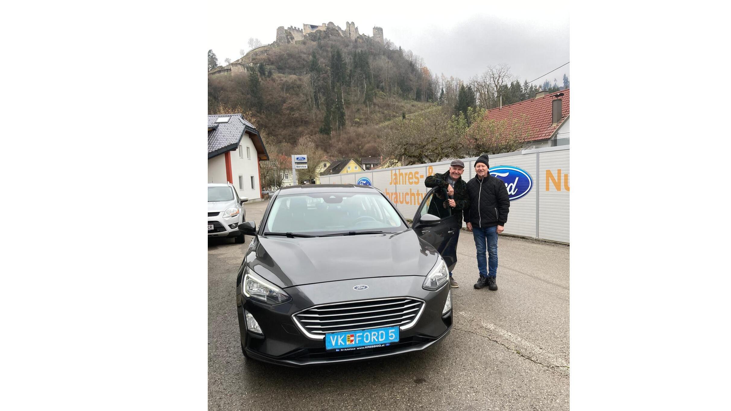 Zwei Männer stehen neben einem grauen Ford in einer Stadt, mit einer Burg im Hintergrund.