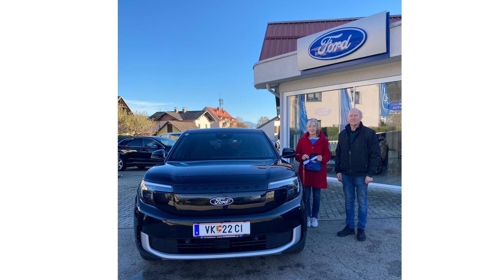 Ein Paar steht neben einem schwarzen Ford-Auto vor einem Ford-Händler. Heller Himmel und Gebäude im Hintergrund.