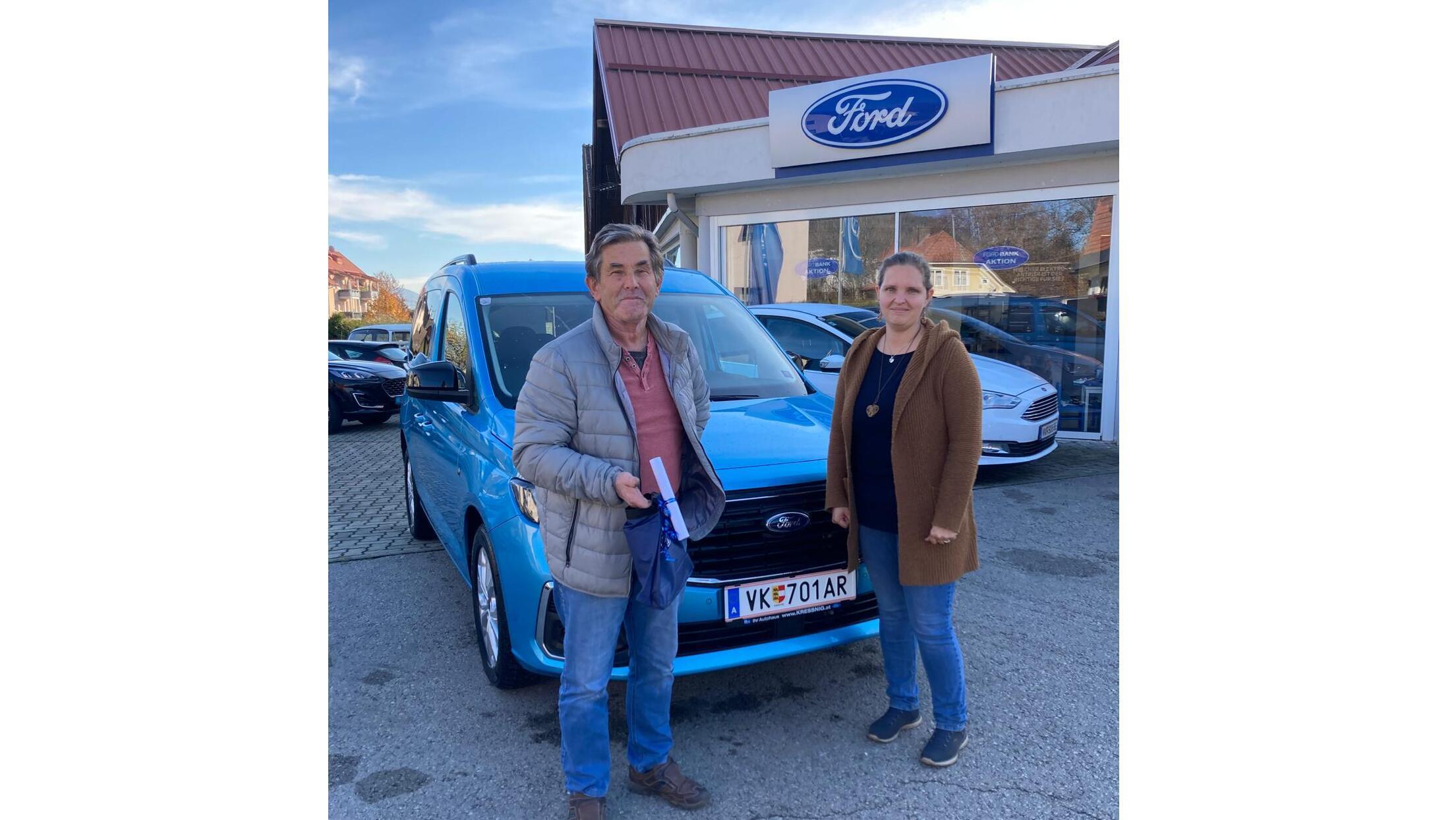 Ein Mann und eine Frau stehen neben einem blauen Ford Auto vor einem Autohaus. Ein sonniger Tag.
