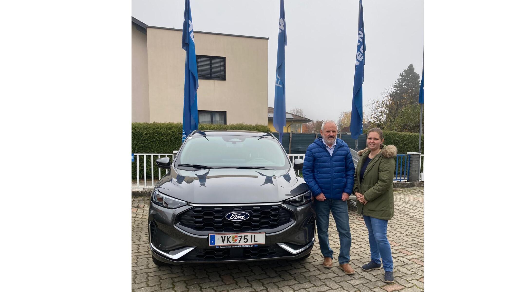 Zwei Personen stehen neben einem schwarzen Ford-Auto mit blauen Fahnen im Hintergrund.