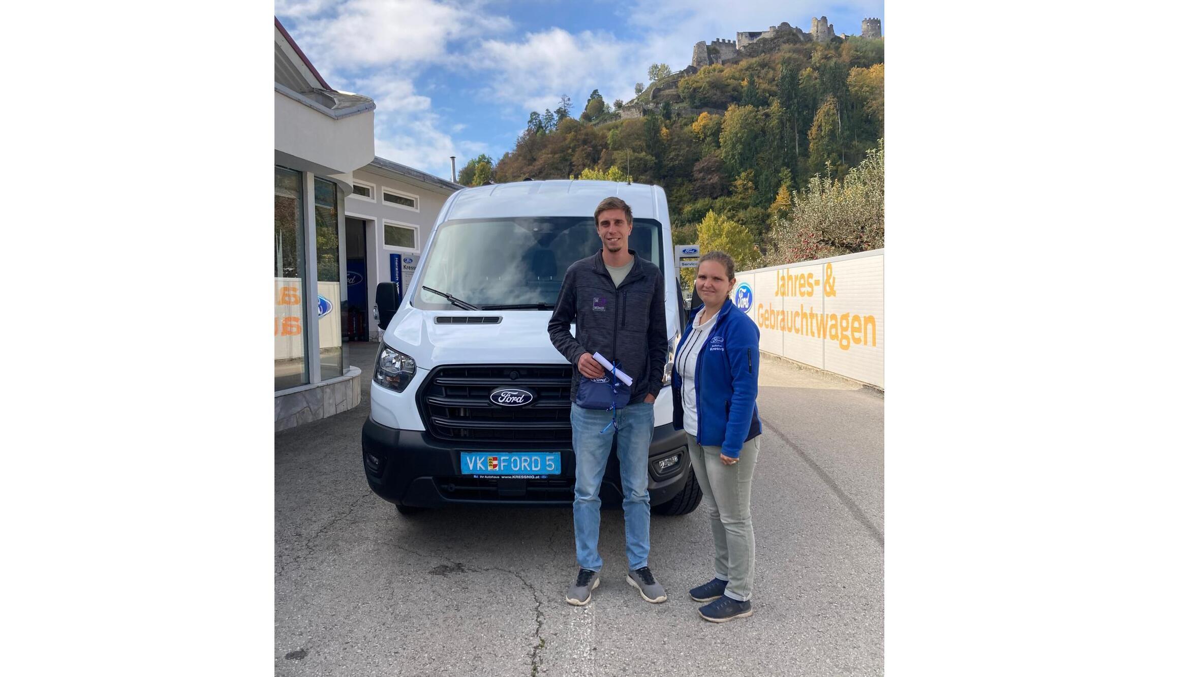 Ein Mann und eine Frau stehen neben einem weißen Ford Transporter vor einer malerischen Landschaft.