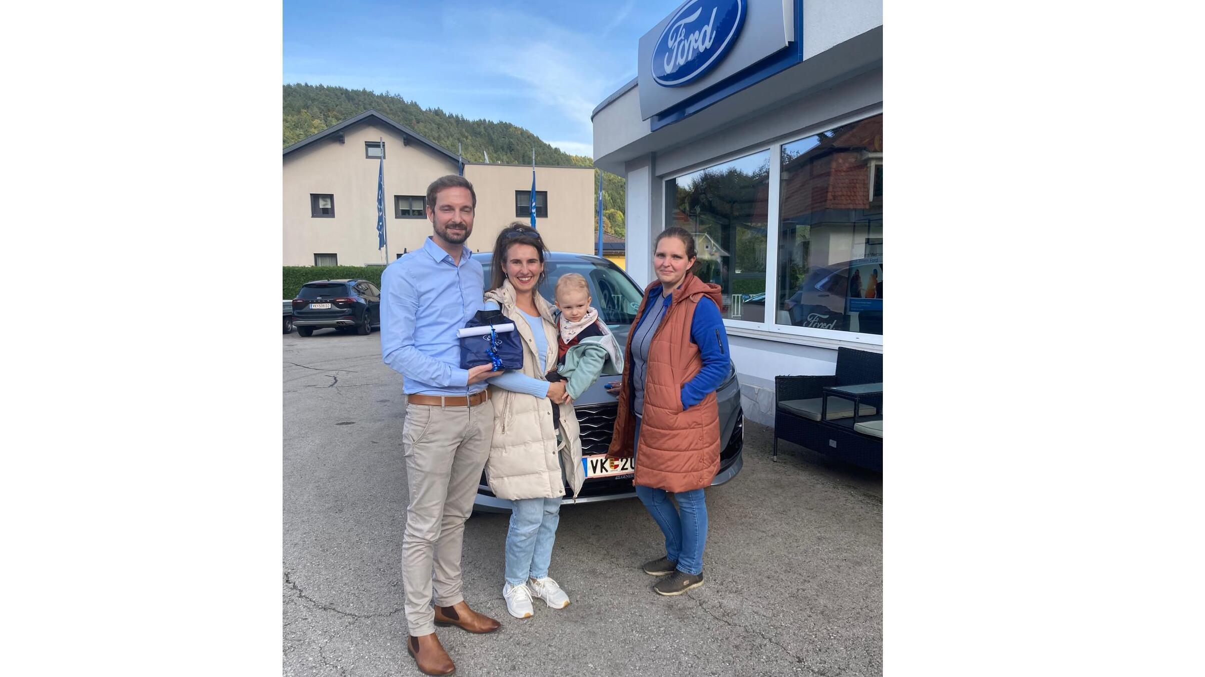 Familie steht vor einem Ford-Autohaus mit einem neuen Auto, gemeinsam mit einem Berater und einem Kind.