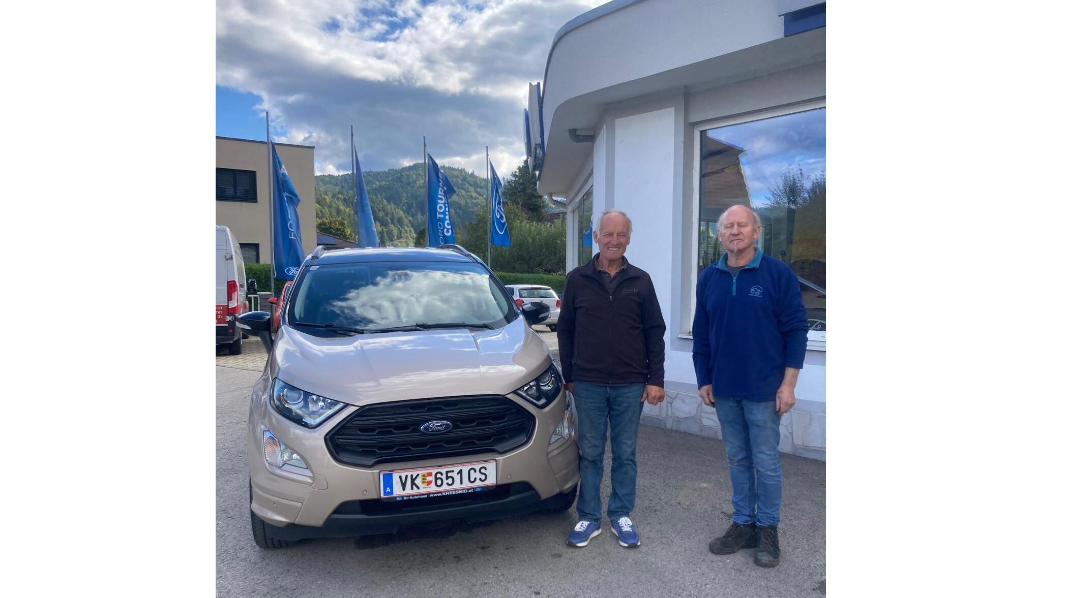 Zwei Männer stehen neben einem beige Ford EcoSport vor einem Autohaus mit blauen Fahnen.