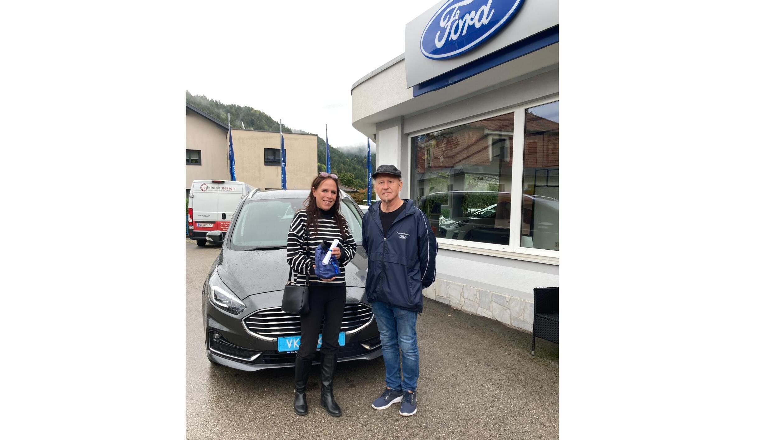 Zwei Personen stehen vor einem Ford-Auto und einem Ford-Händler, strahlende Gesichter und ein neuer Fahrzeugschlüssel.