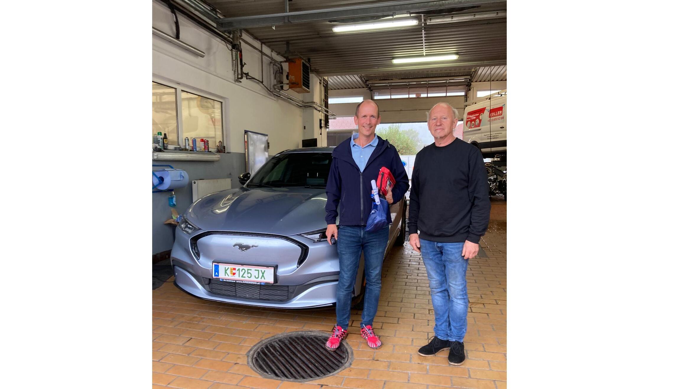 Zwei Männer stehen vor einem grauen Auto in einer Werkstatt, umgeben von Werkzeug und Ausrüstung.