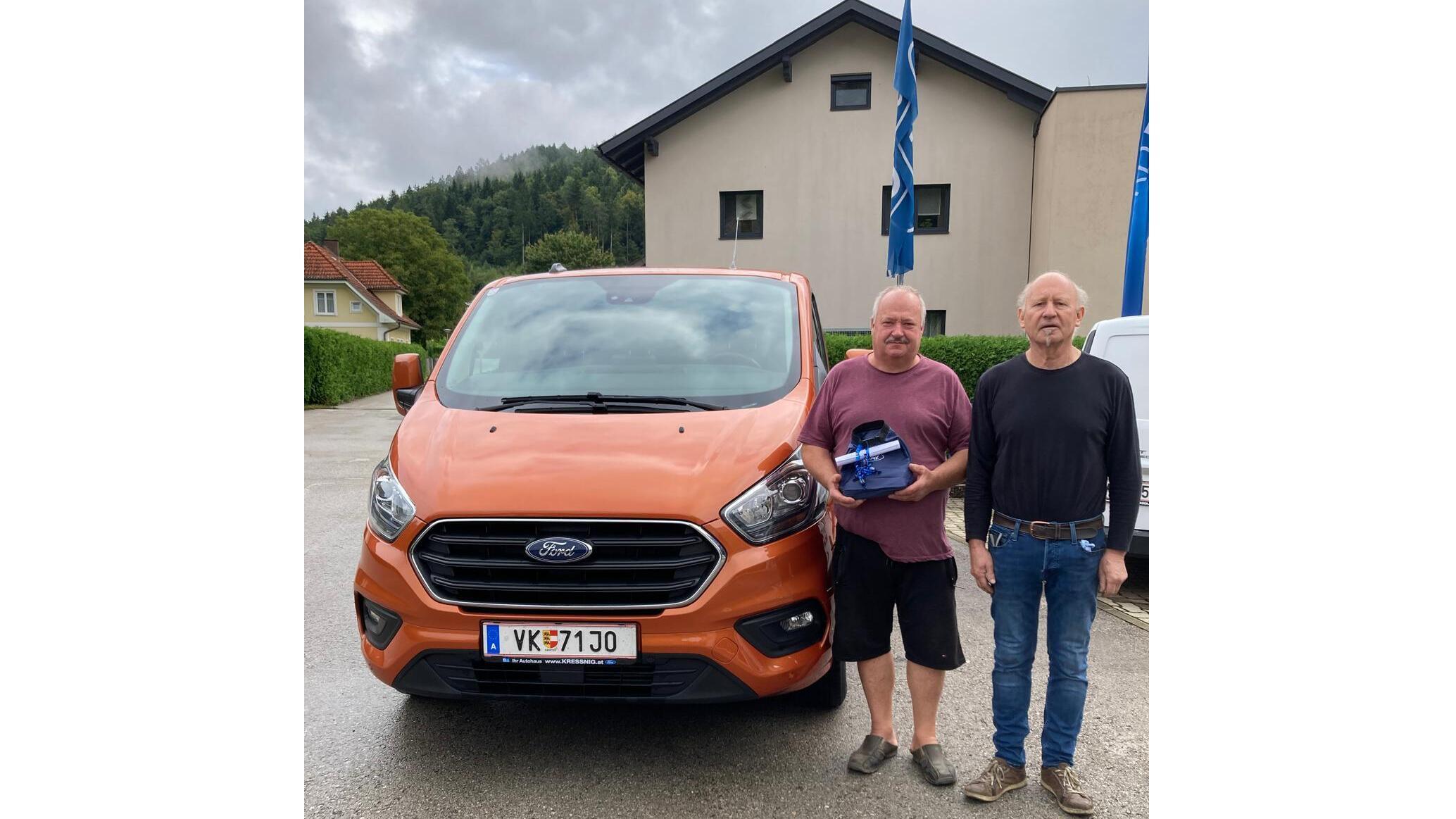 Zwei Männer stehen neben einem orangefarbenen Ford Transporter vor einem Haus in der Natur.