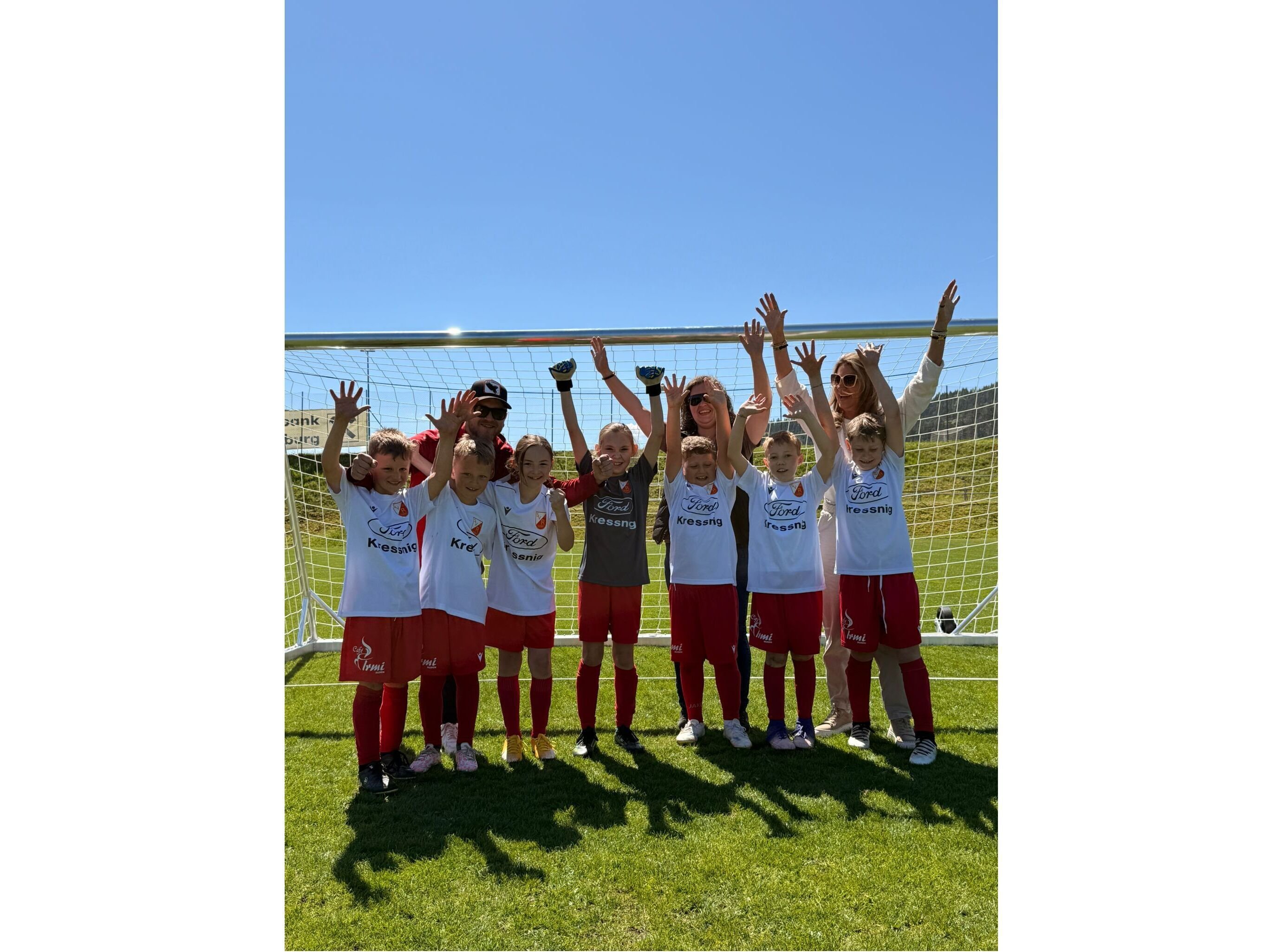 Eine Gruppe von Kindern und Erwachsenen in Fußballtrikots posiert fröhlich vor einem Tor bei strahlendem Sonnenschein.