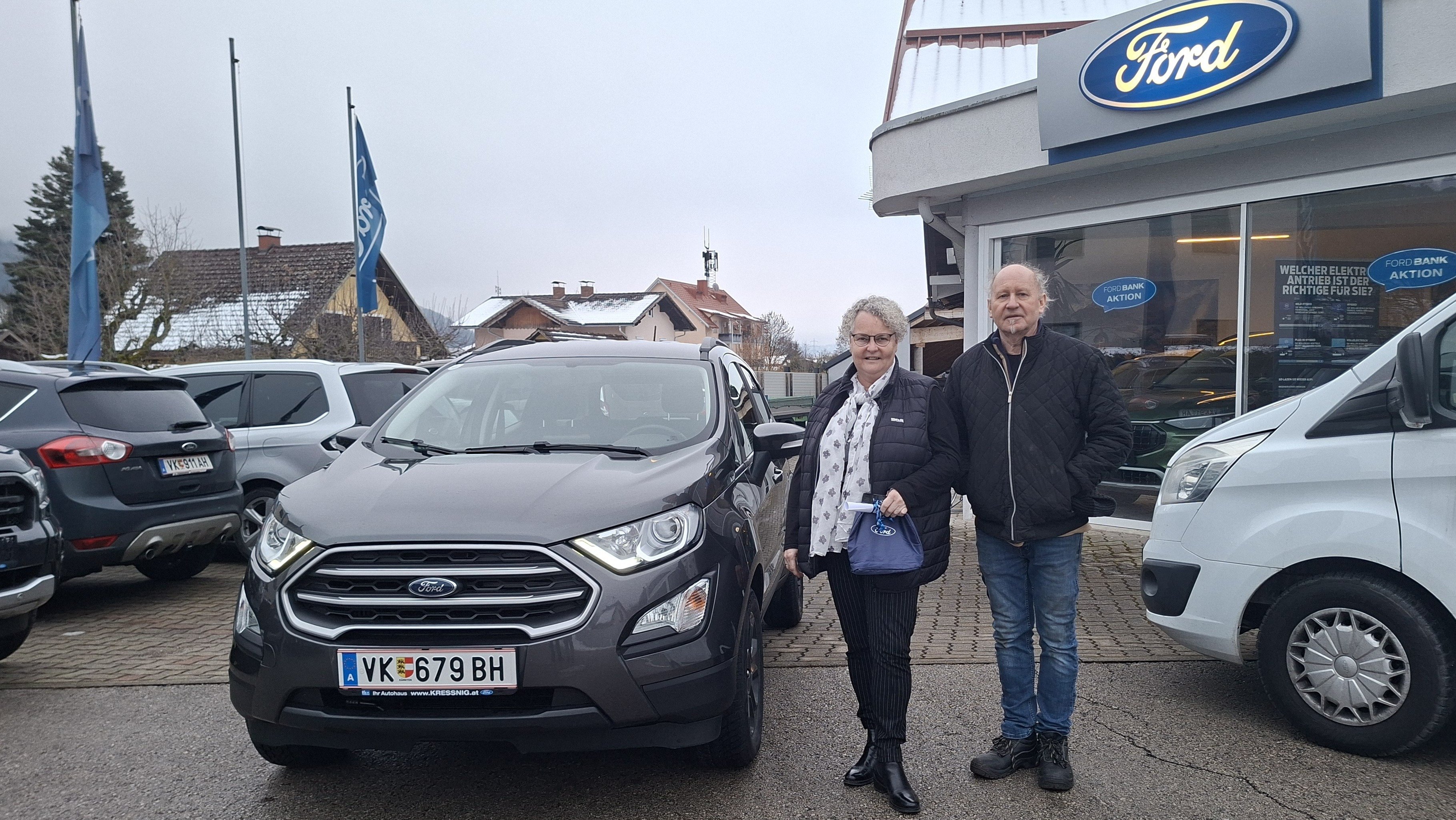 Ein älteres Paar steht neben einem grauen Ford EcoSport auf einem Autohausgelände mit Ford-Flagge im Hintergrund.