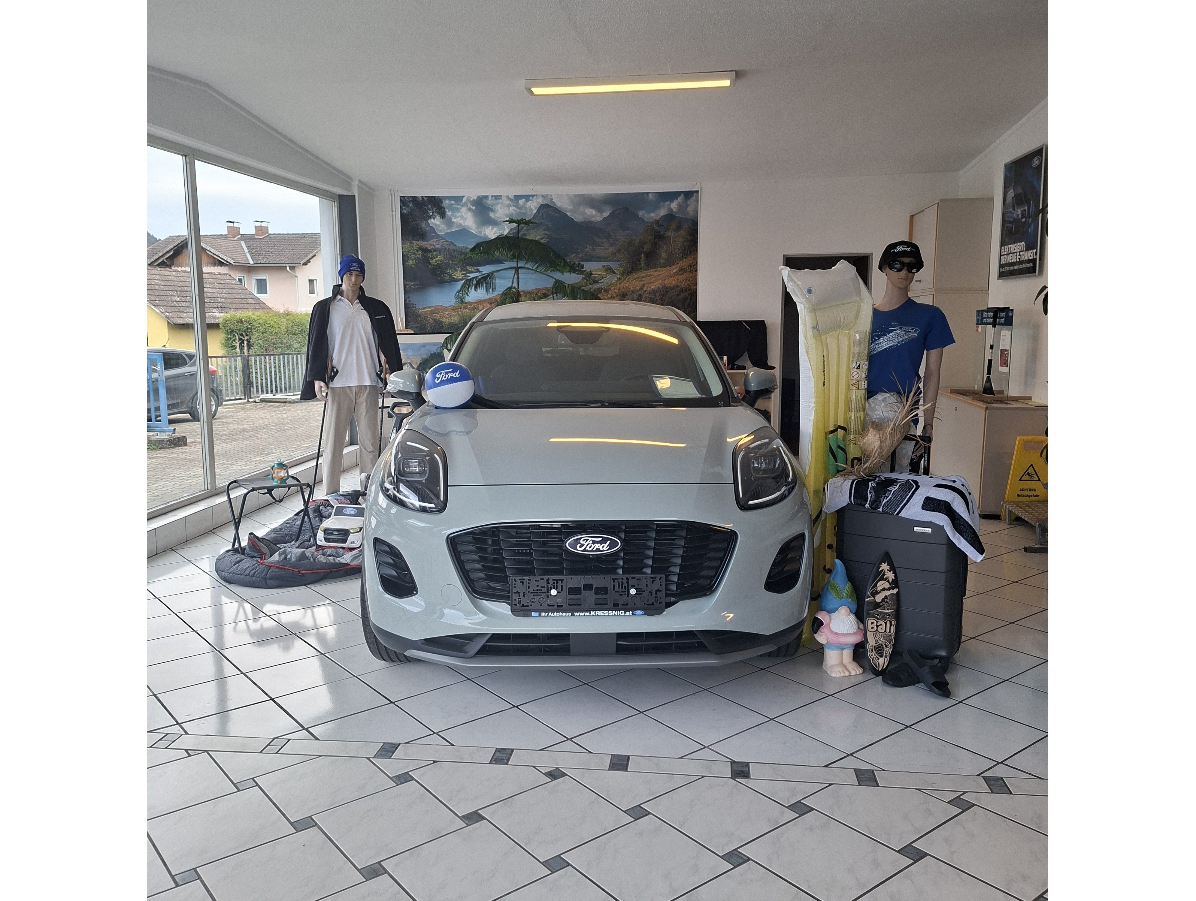 Ein grauer Ford PKW steht in einem Ausstellungsraum, umgeben von Sportutensilien und Schaufensterpuppen.