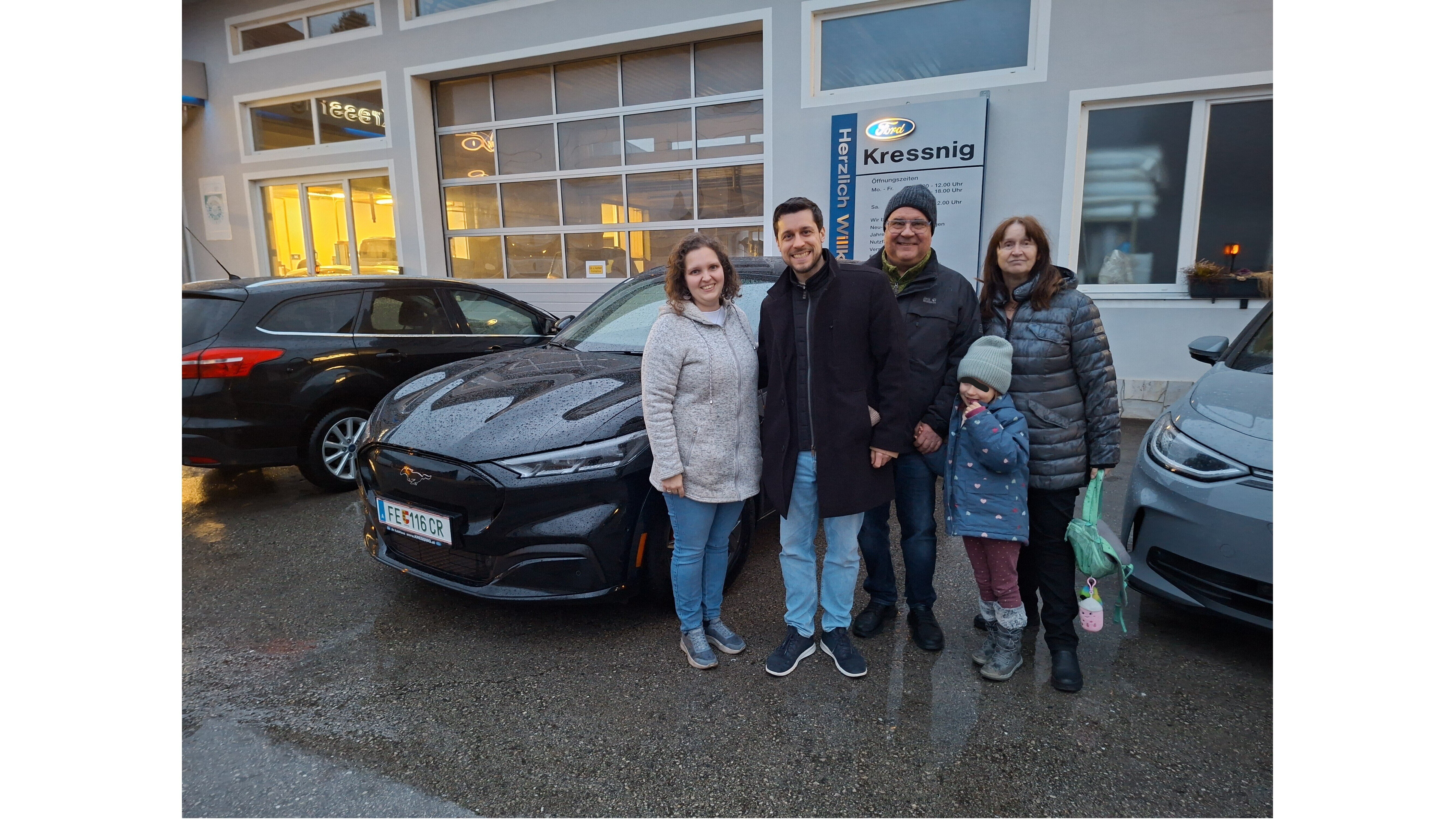 Familie steht neben einem schwarzen Auto vor einem Autohaus, bei kaltem Wetter mit Neuschnee.
