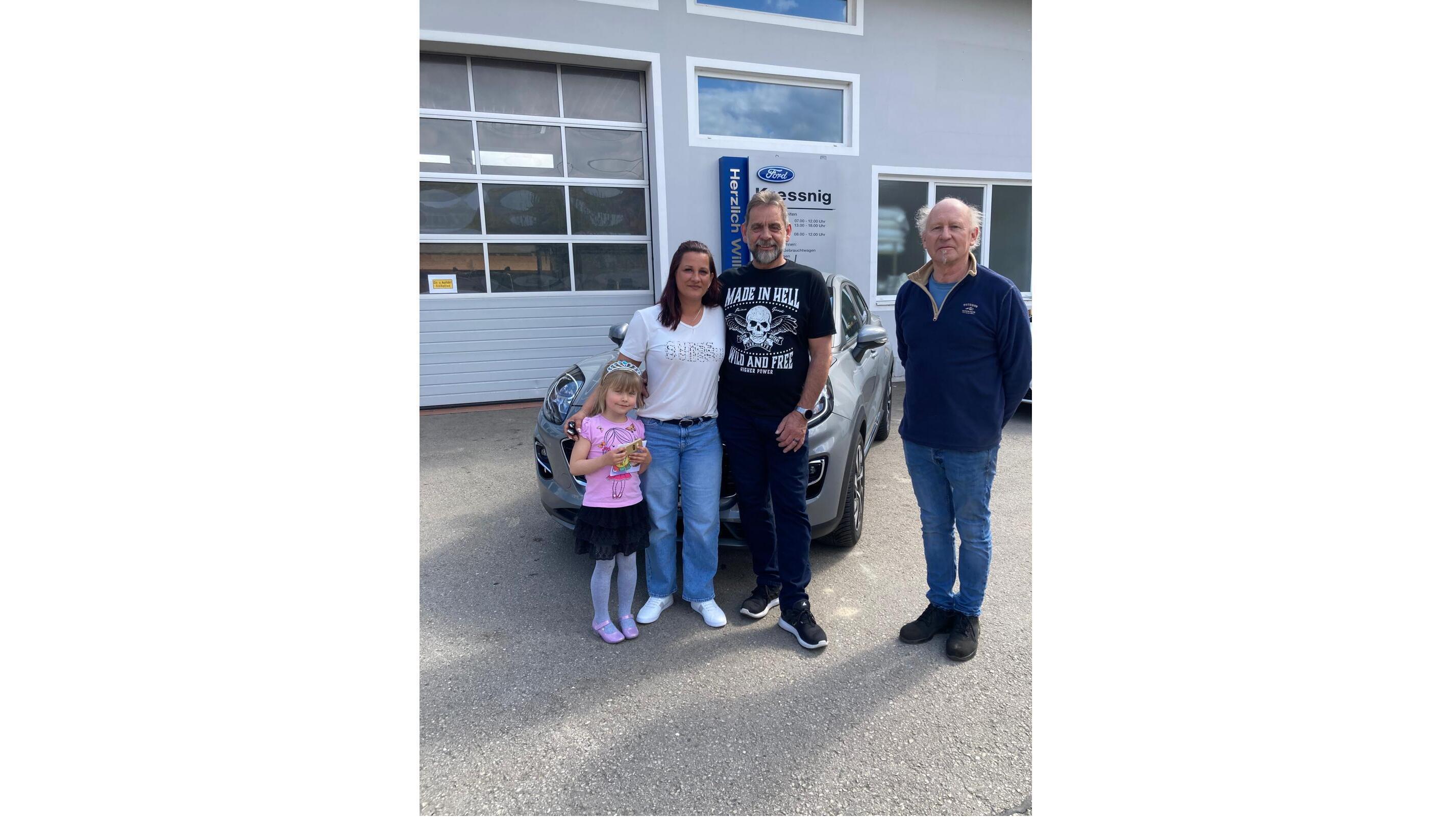Eine Familie steht vor einem Autohaus mit einem neuen Auto. Vier Personen, darunter ein Kind, lächeln in die Kamera.
