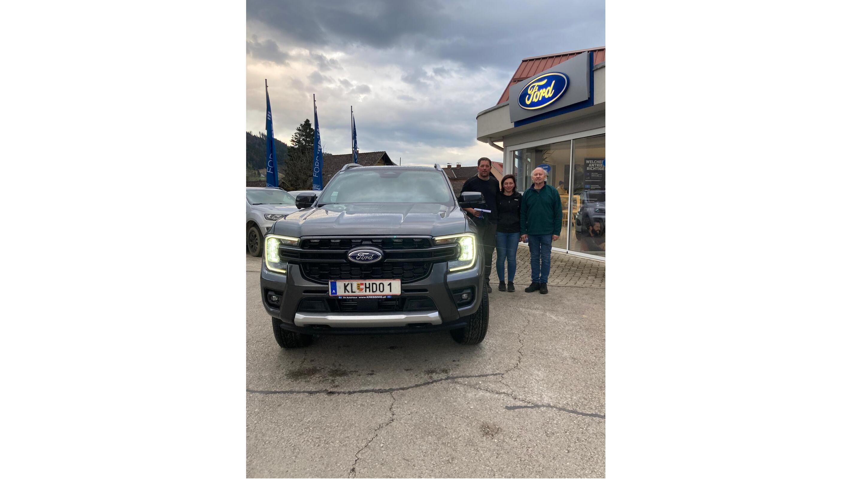 Drei Personen stehen neben einem neuen Ford-Truck vor einem Ford-Händler. Wolkiger Himmel im Hintergrund.
