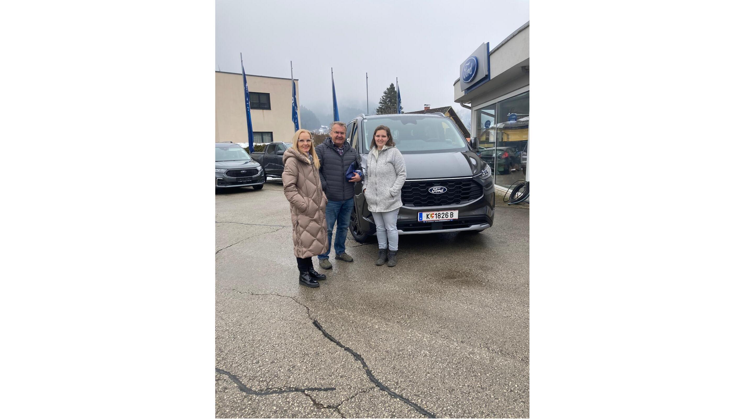 Drei Personen stehen vor einem neuen Ford im Autohaus, umgeben von Fahrzeugen und Fahnen.