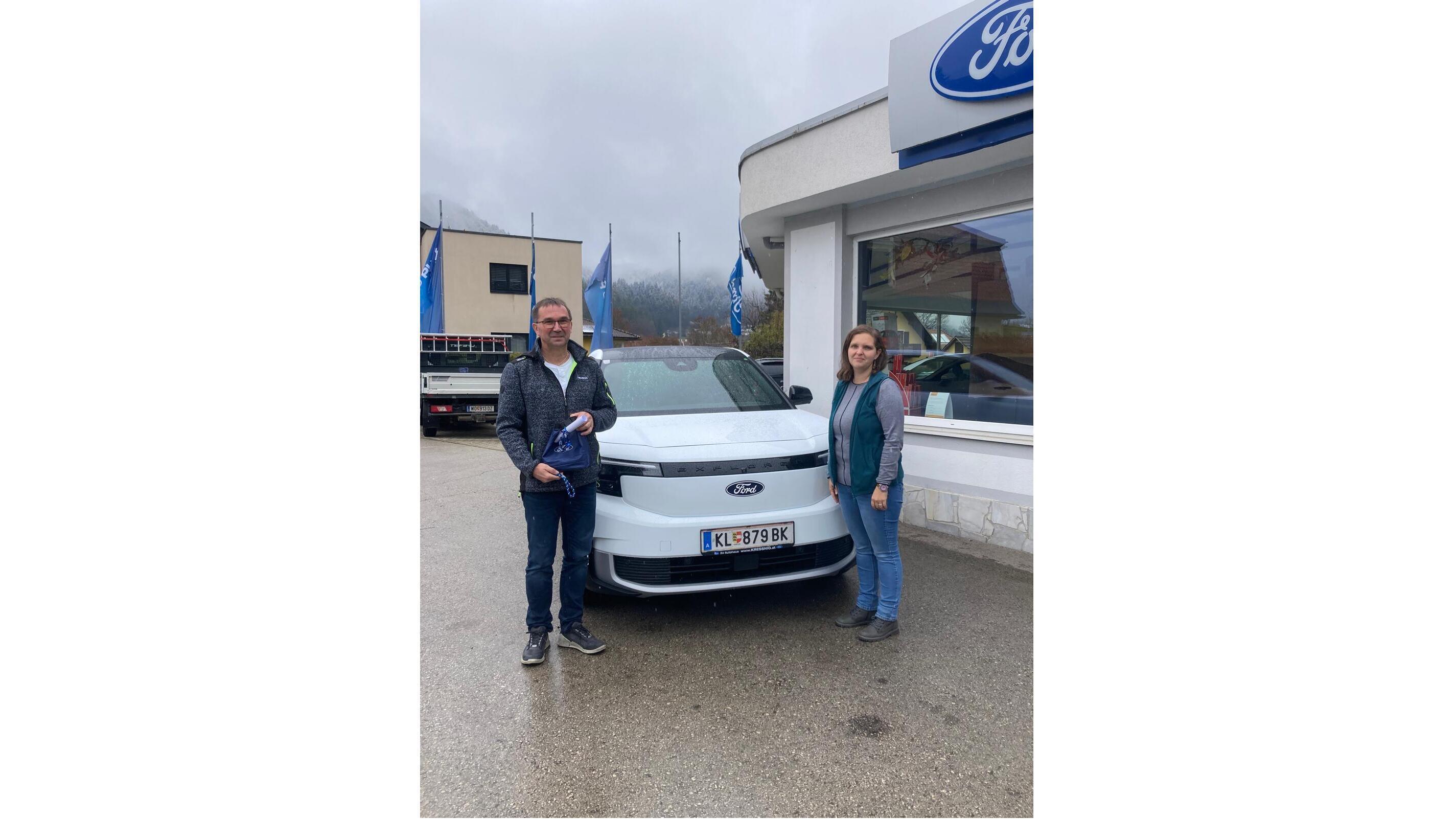 Zwei Personen stehen neben einem weißen Ford Auto vor einem Autohaus, bewölkter Himmel im Hintergrund.