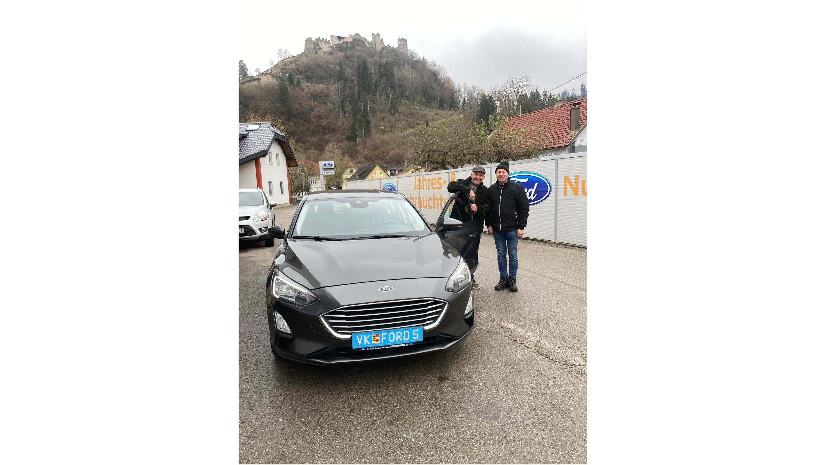 Zwei Männer stehen neben einem grauen Ford-Fahrzeug mit Burg im Hintergrund.