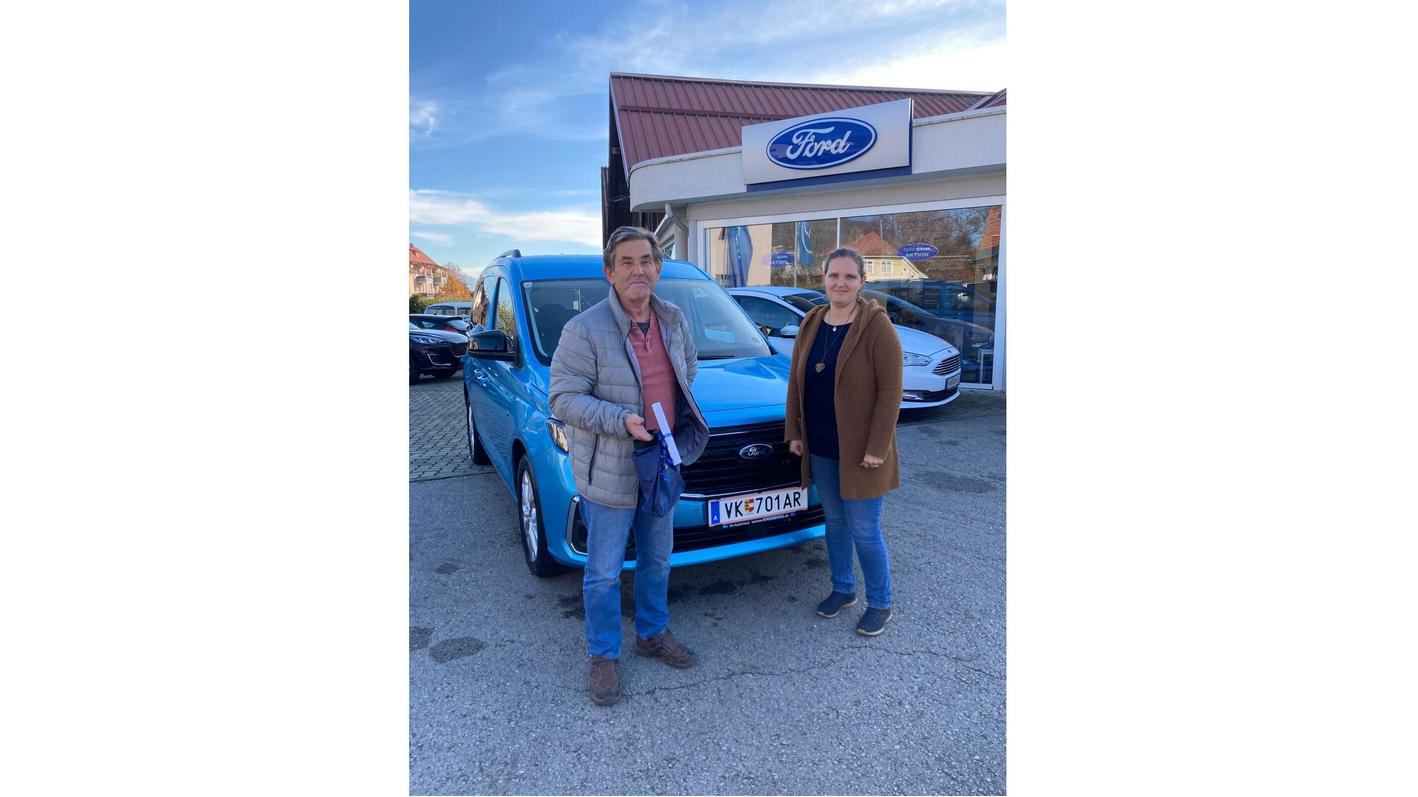 Ein Mann und eine Frau stehen vor einem blauen Ford-Auto in einem Autohaus, lächelnd und bereit für einen Kauf.
