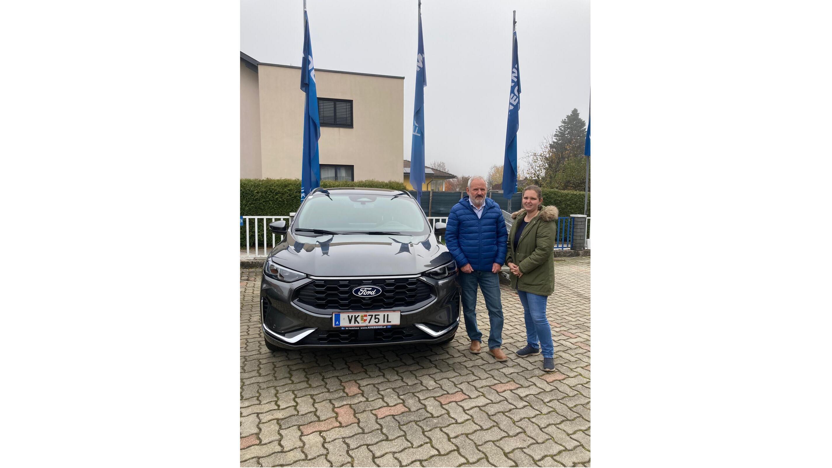 Zwei Personen stehen neben einem schwarzen Ford-Auto, umgeben von Flaggen vor einem Gebäude.