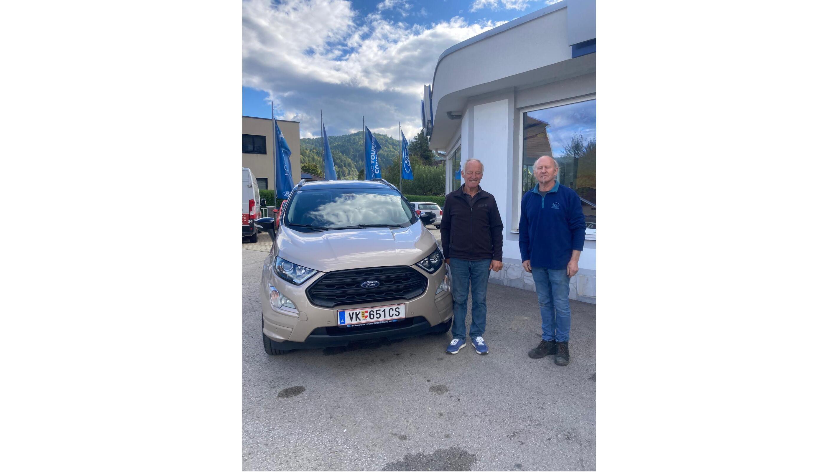 Zwei Männer stehen neben einem neuen, beigen Ford Ecosport vor einem Autohaus mit blauen Fahnen.
