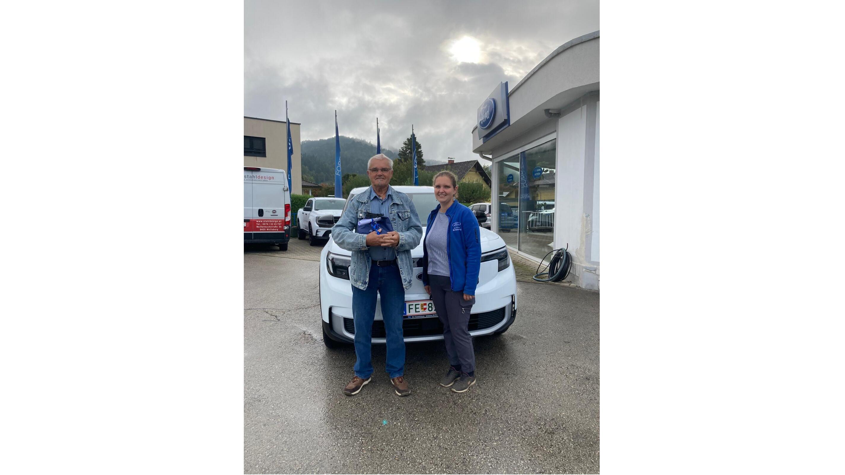 Zwei Personen stehen vor einem weißen Auto in einem Autohaus, Berge im Hintergrund, leicht bewölkter Himmel.