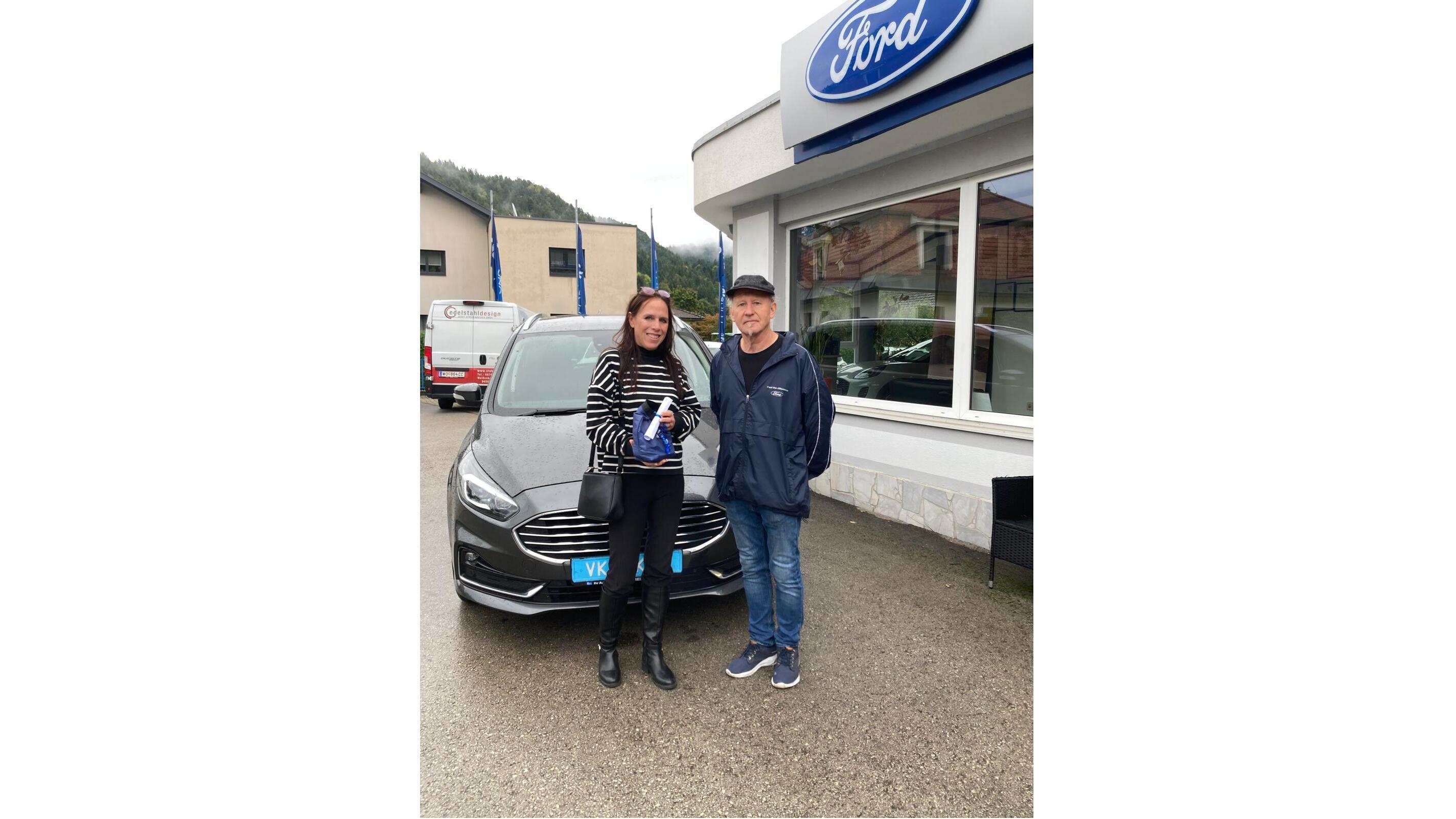 Zwei Personen stehen vor einem neuen Ford-Auto in einem Autohaus, lächelnd und posierend für das Foto.