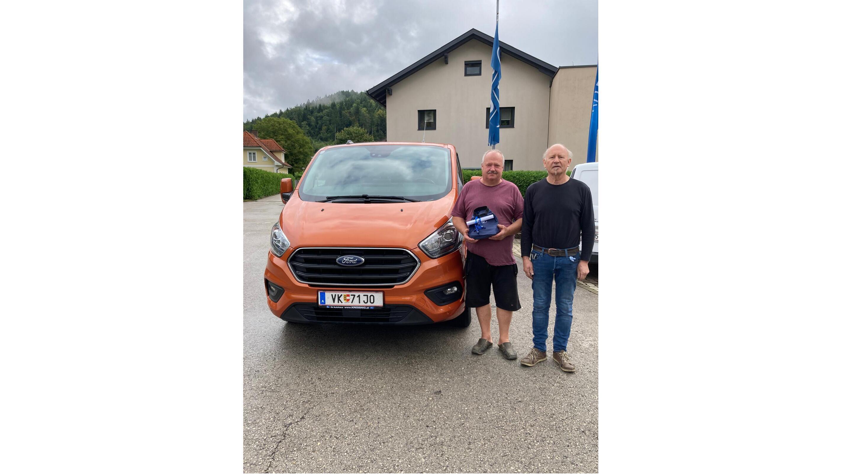 Zwei Männer stehen neben einem orangefarbenen Ford Transit vor einem Gebäude in der Natur.