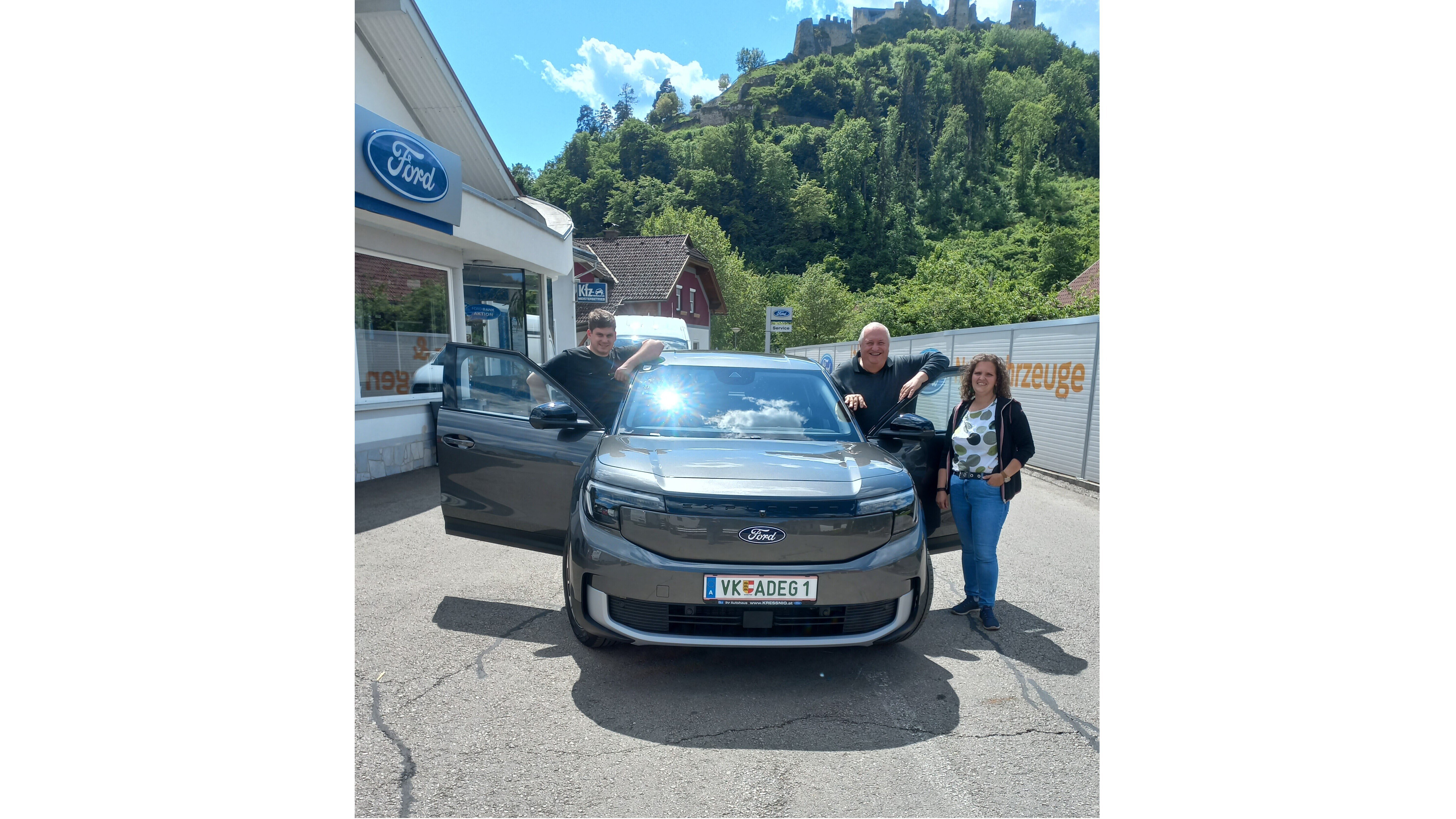 Drei Personen stehen um ein graues Ford-Fahrzeug, vor einem Autohaus in einer malerischen Landschaft.