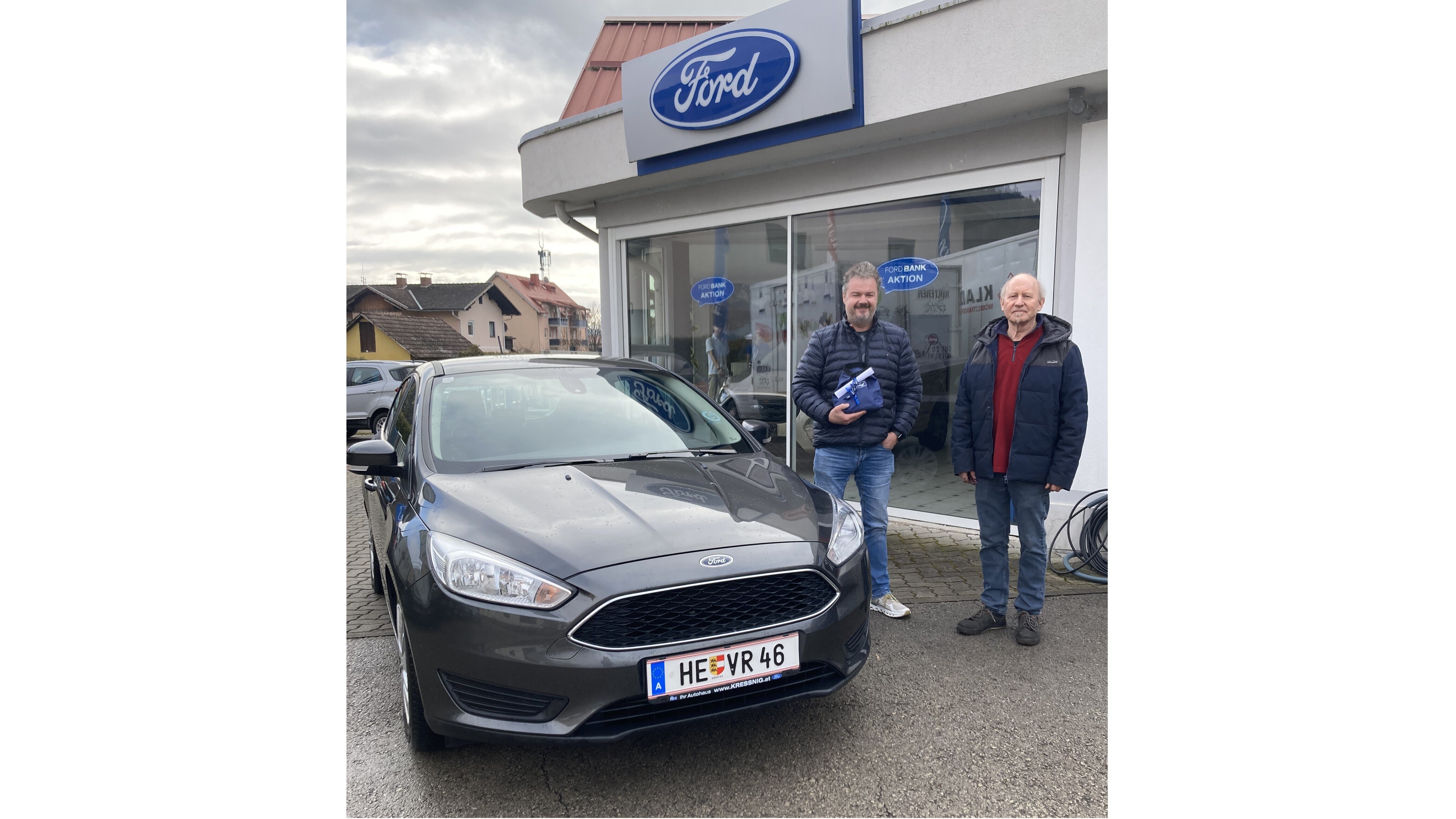 Zwei Männer stehen neben einem grauen Ford vor einem Ford-Autohaus. Das Bild zeigt die Fahrzeugübergabe.