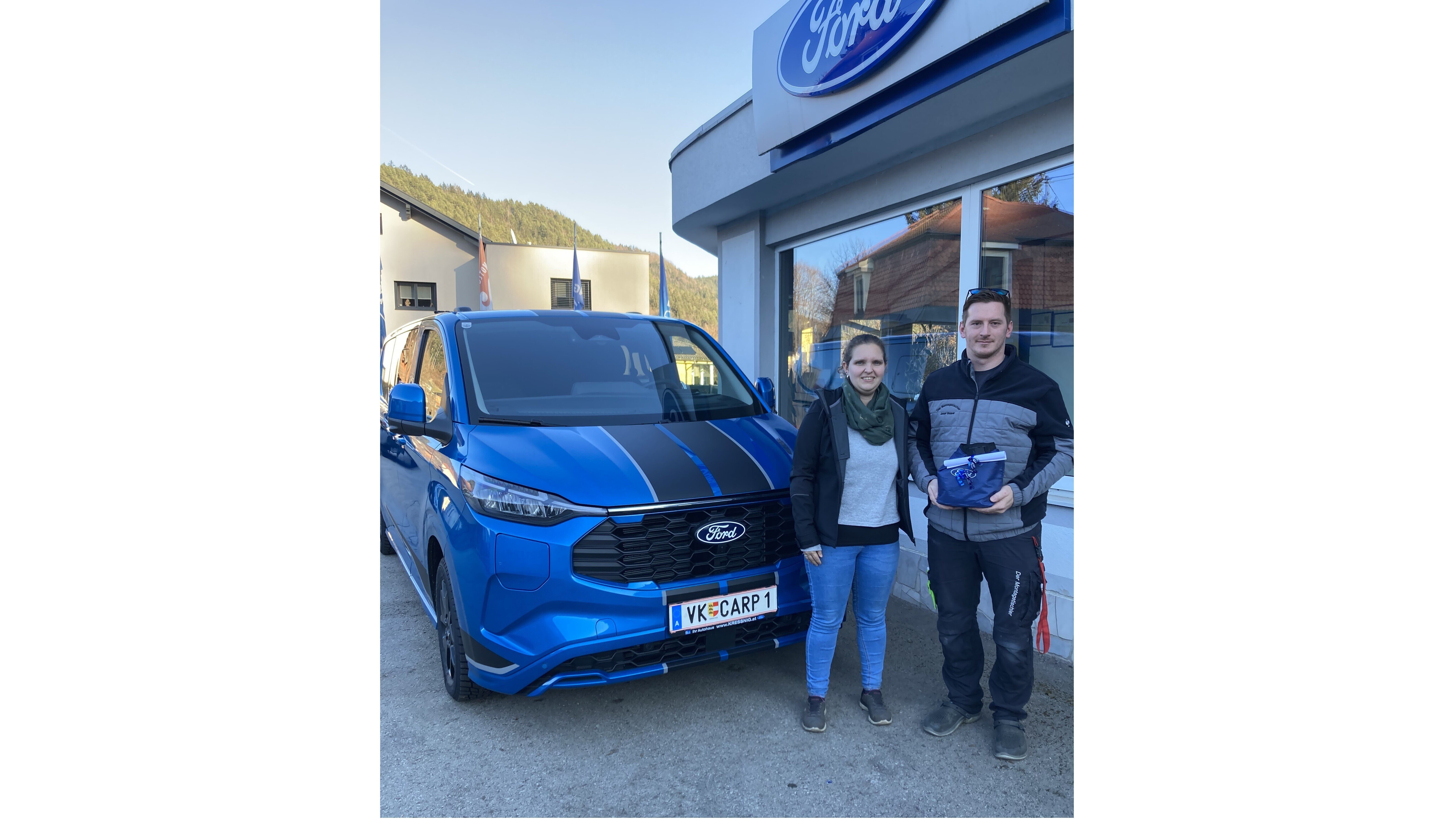 Zwei Personen stehen neben einem blauen Ford vor einem Autohaus. Sie halten einen Schlüssel in der Hand.
