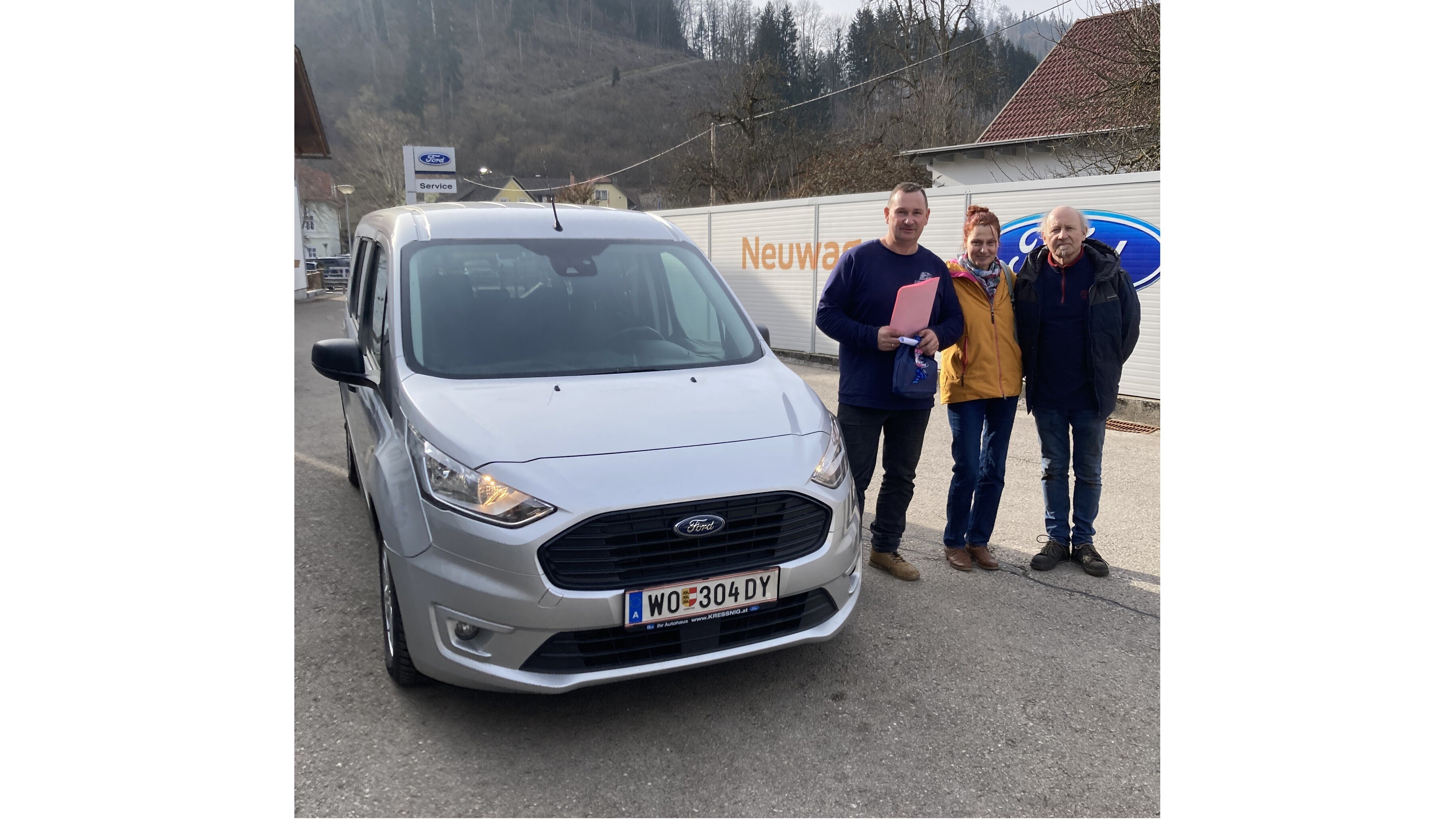 Eine graue Ford-Fahrzeug steht in der Nähe eines Autohauses. Drei Personen posieren mit Dokumenten.