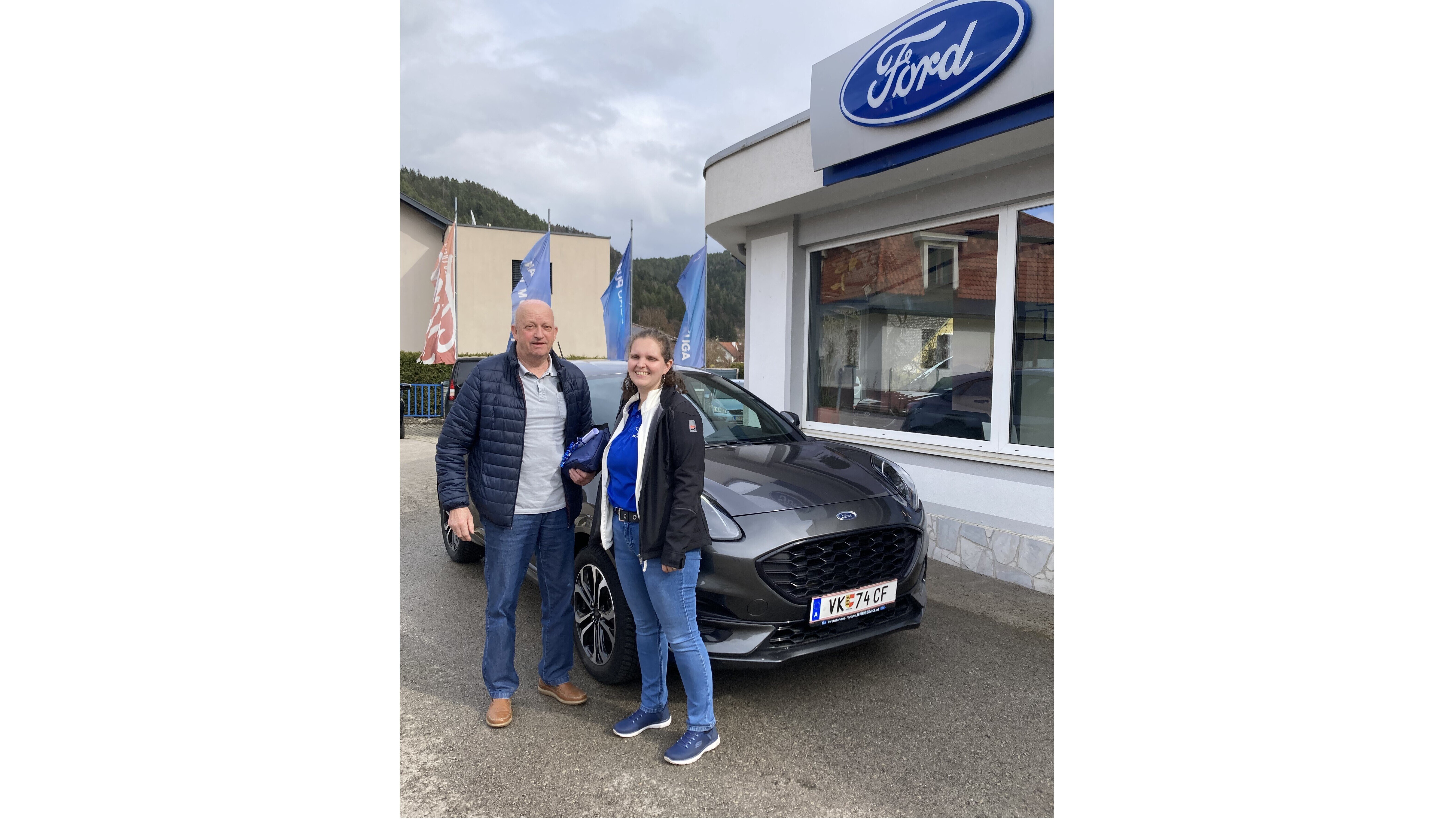 Ein Mann und eine Frau stehen vor einem grauen Ford-Auto neben einem Ford-Händler. Sie lächeln und halten den Autoschlüssel.