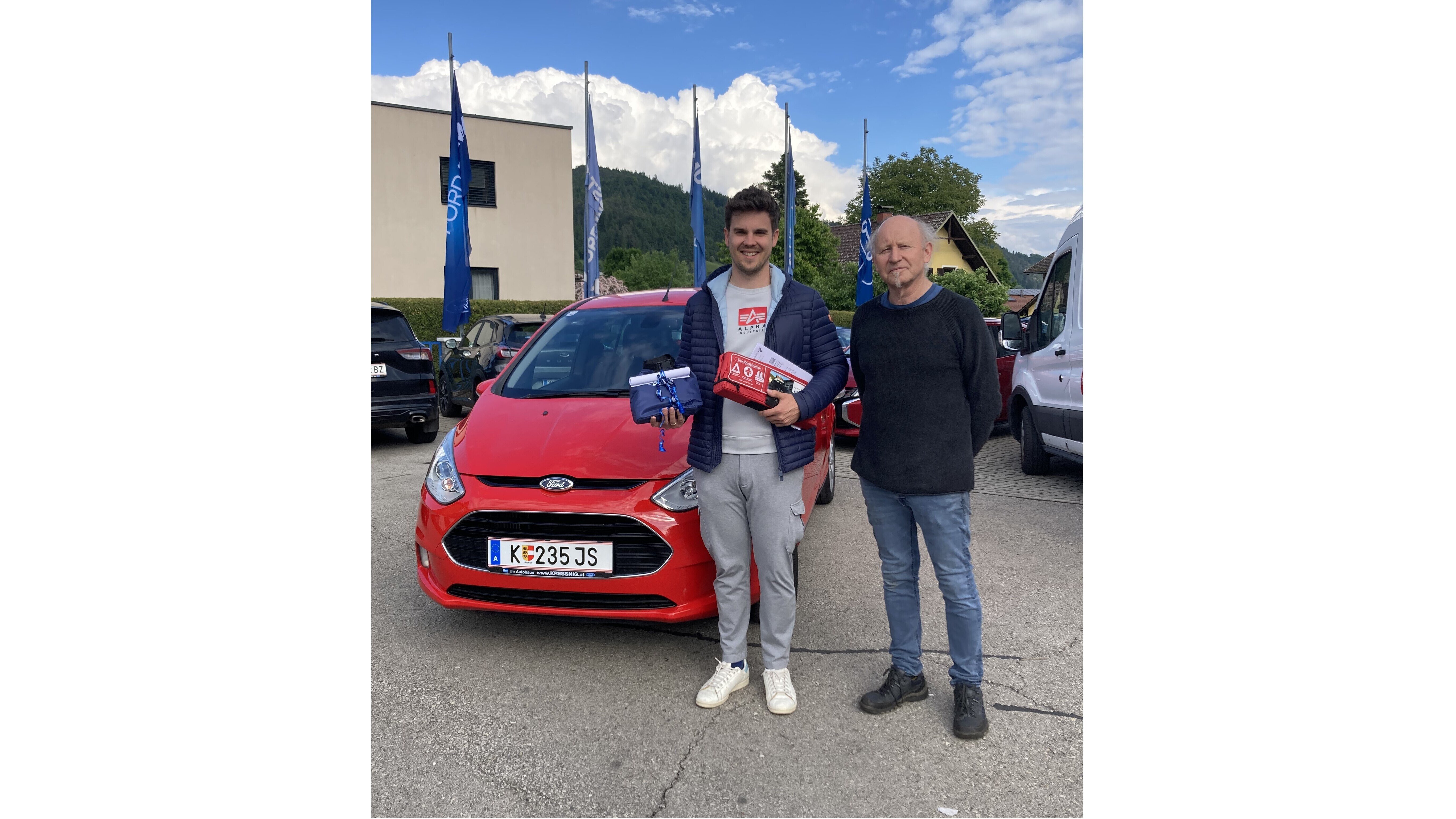 Zwei Männer stehen vor einem roten Ford-Auto und halten Geschenke in der Hand. Im Hintergrund sind Bäume und Gebäude sichtbar.