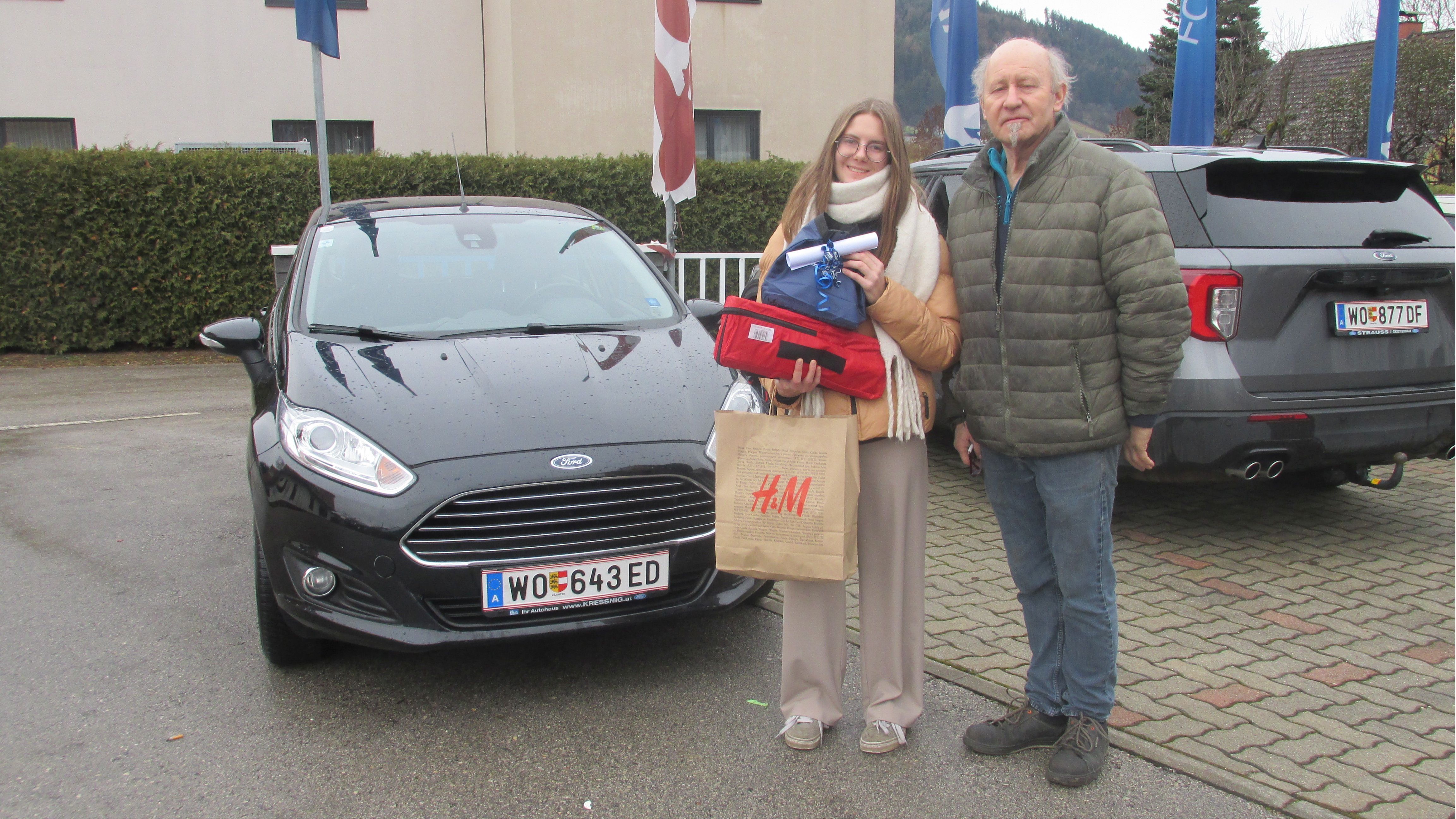 Eine Frau steht mit einem Mann neben einem schwarzen Ford Fiesta und hält Einkaufstaschen in der Hand.