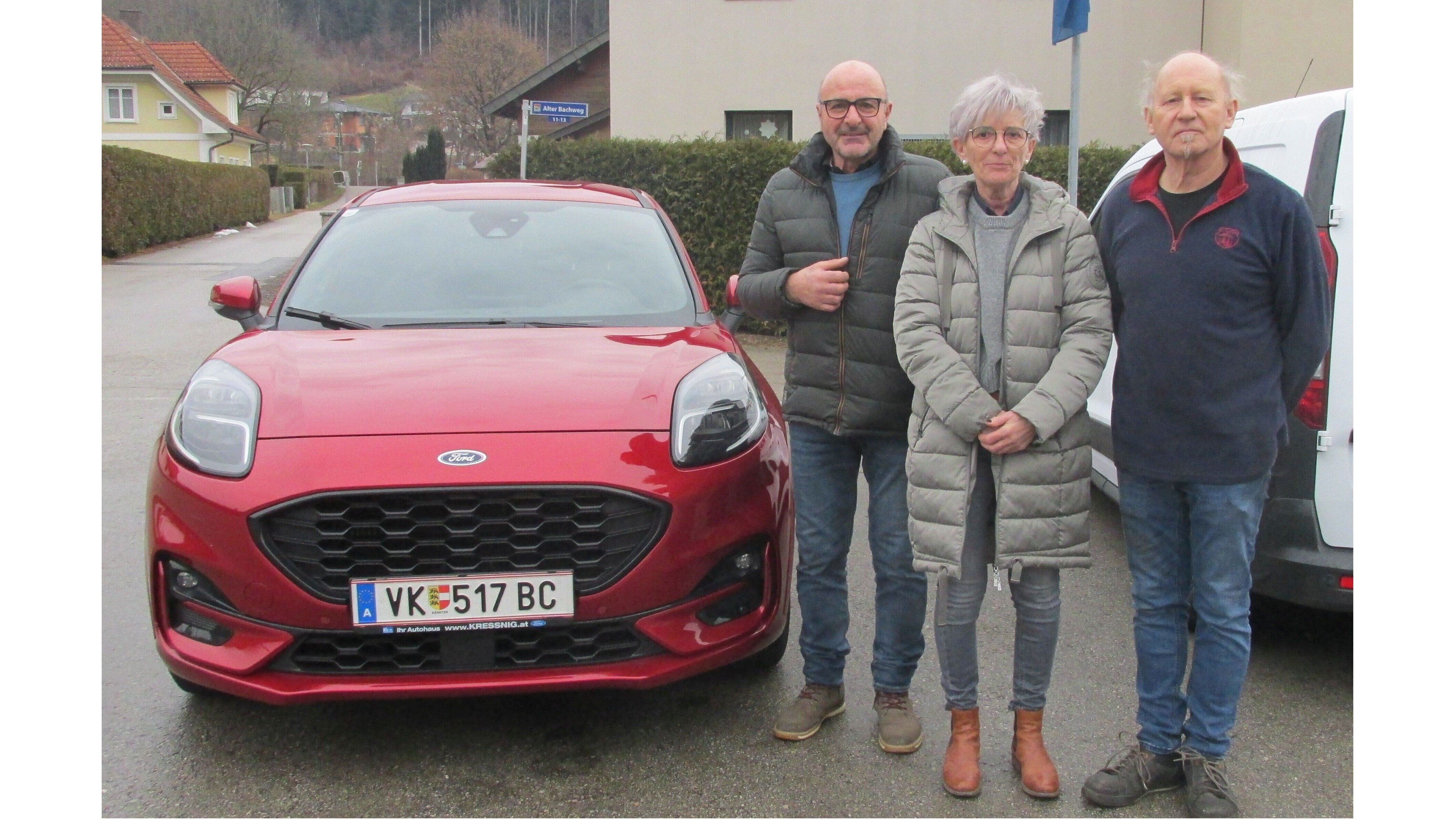 Drei Personen stehen neben einem roten Ford Puma auf einer Straße in einer ländlichen Umgebung.