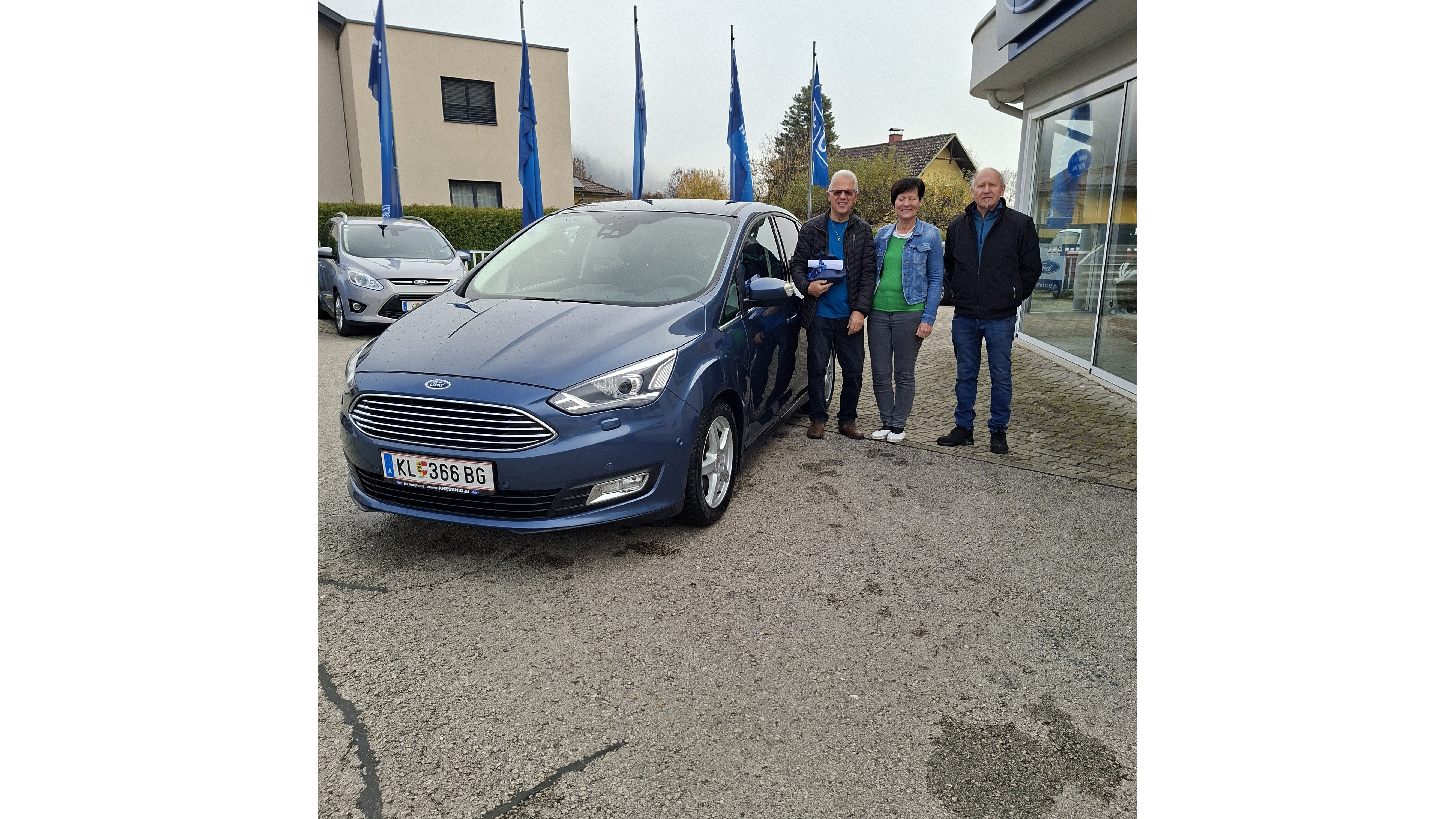 Ein blauer Ford steht vor einem Autohaus, umgeben von vier Personen und Flaggen im Hintergrund.