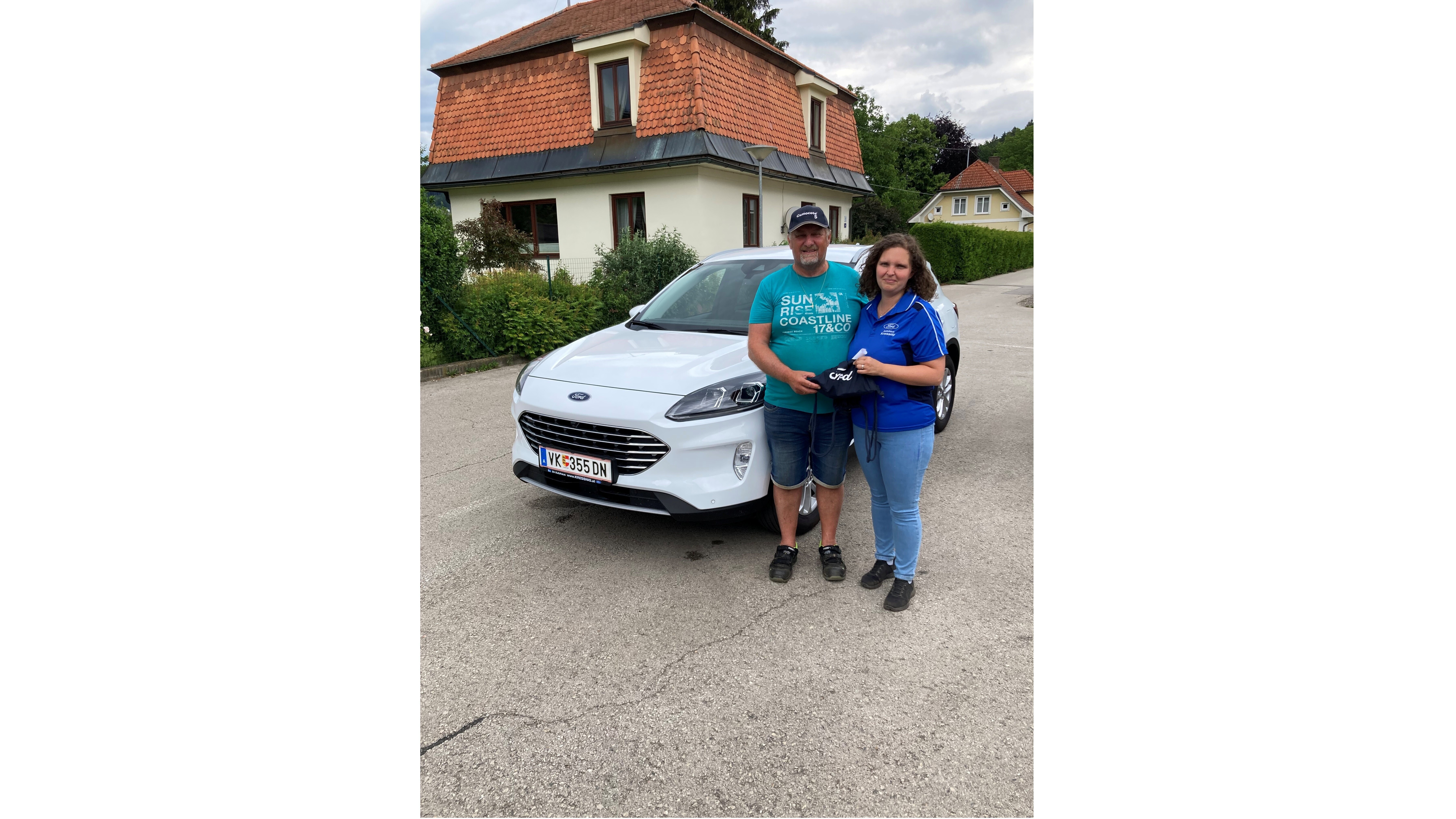 Ein Mann und eine Frau stehen vor einem weißen Ford auf einer Straße, umgeben von Wohnhäusern und Bäumen.