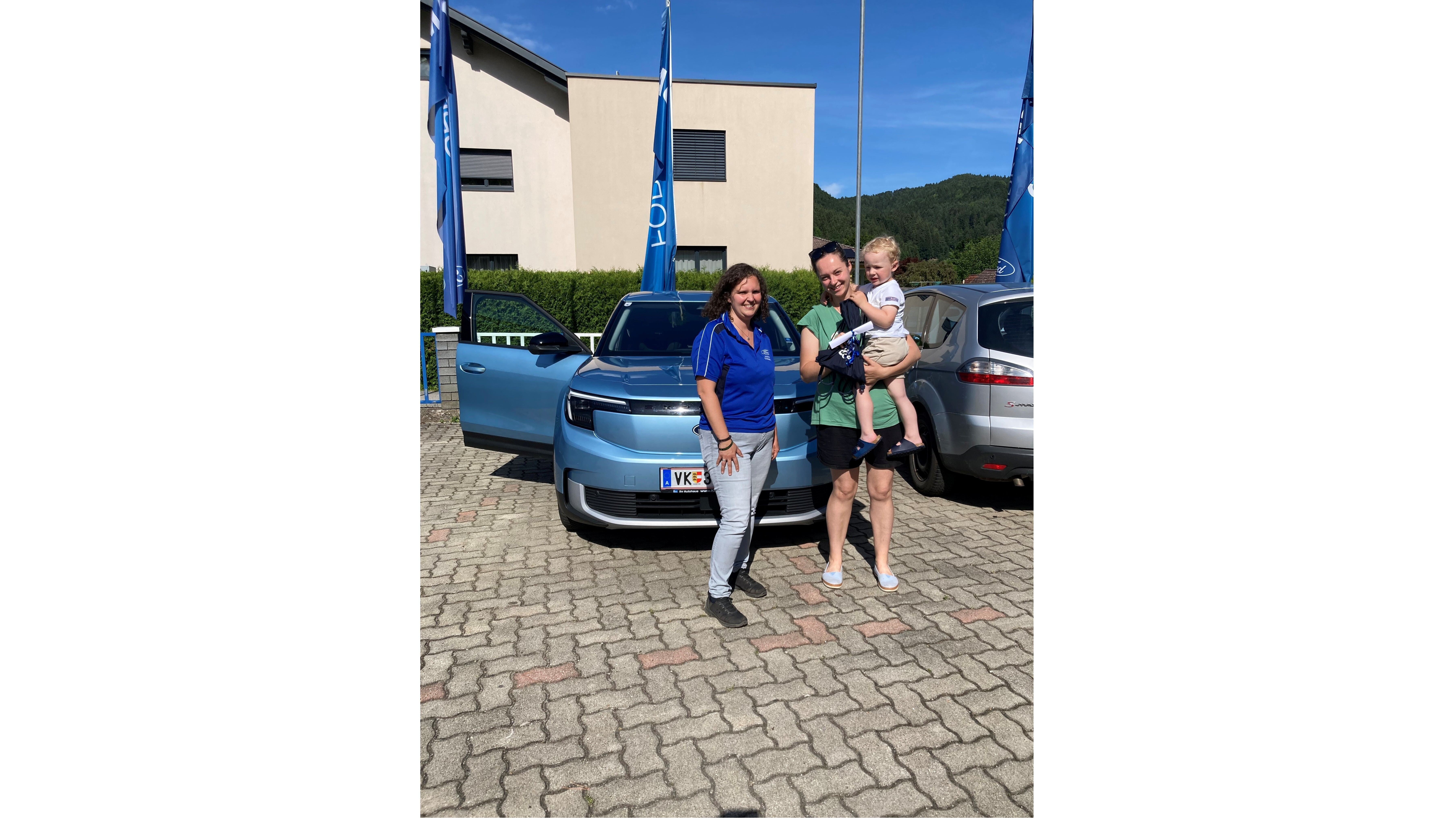 Familie mit einem kleinen Kind steht neben einem blauen Auto auf einem Parkplatz bei sonnigem Wetter.